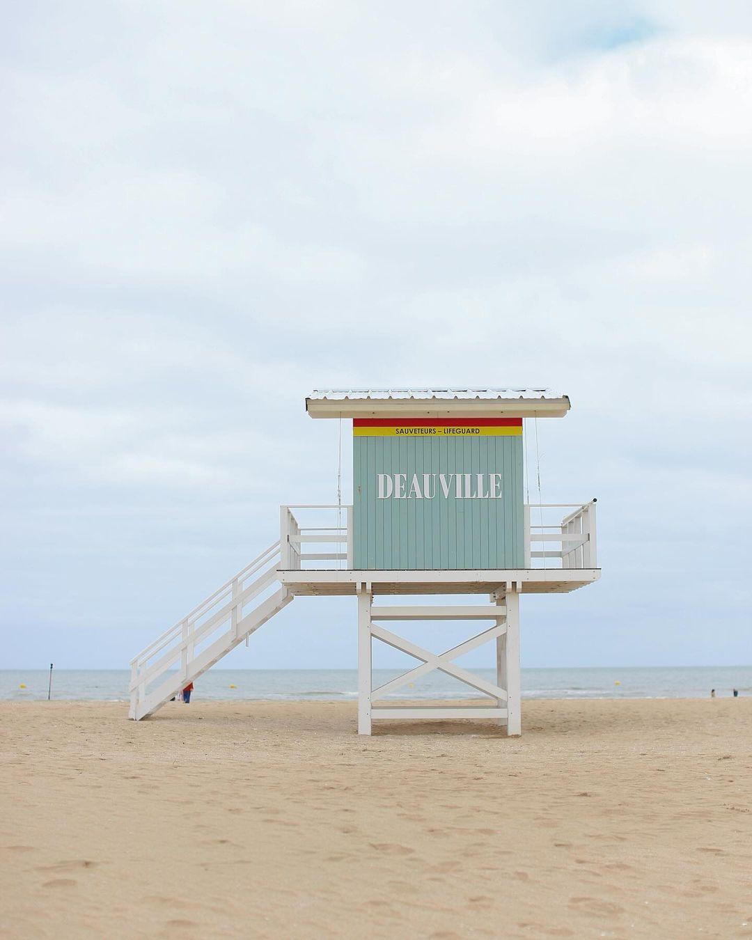 Image of a pastel-colored lifeguard tower on a sandy beach under a cloudy sky. The tower has the word "DEAUVILLE" written on it in large letters, and "Baignade Surveillée" above in smaller yellow text. The ocean is visible in the background.