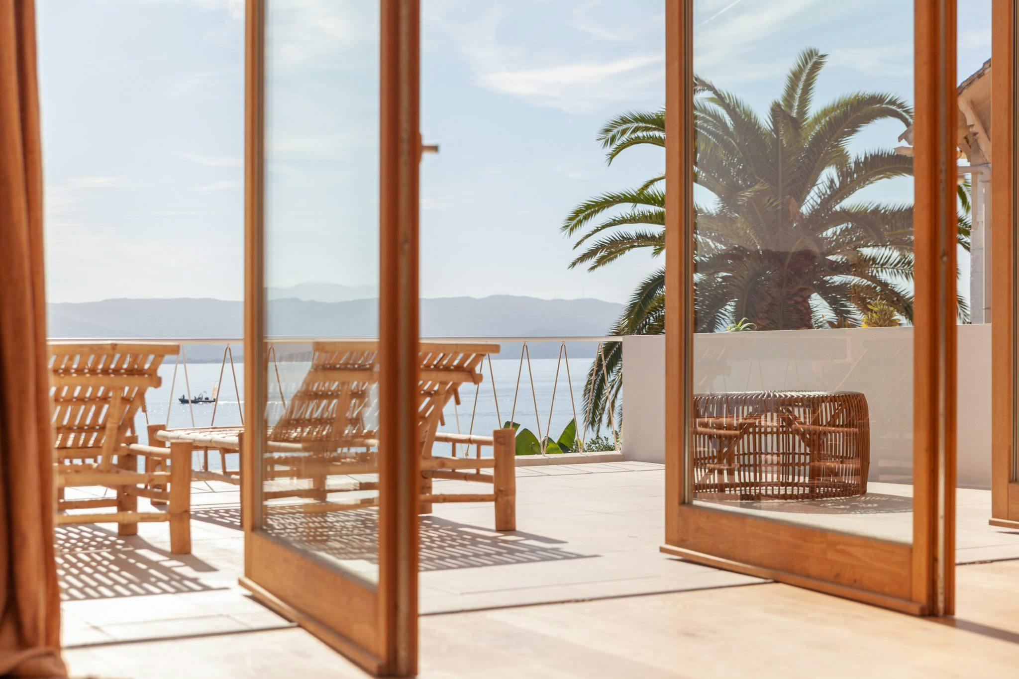 Une terrasse ensoleillée avec des meubles en bois, dont des chaises et des tables, est visible à travers des portes vitrées ouvertes. En arrière-plan, il y a un palmier, un ciel bleu clair et une vue sur l'océan avec des montagnes au loin. Cette scène sereine rappelle des destinations de luxe comme la Corse.