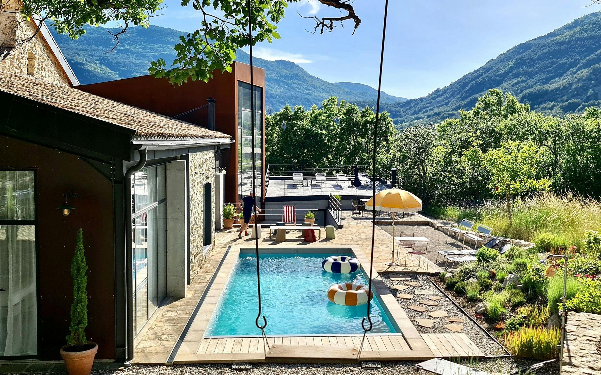 A serene backyard scene featuring a swimming pool surrounded by a deck, loungers, and a swing. The pool has two inflatable rings. In the background, there are mountains and lush greenery under a clear sky. A modern house is adjacent to the pool area.