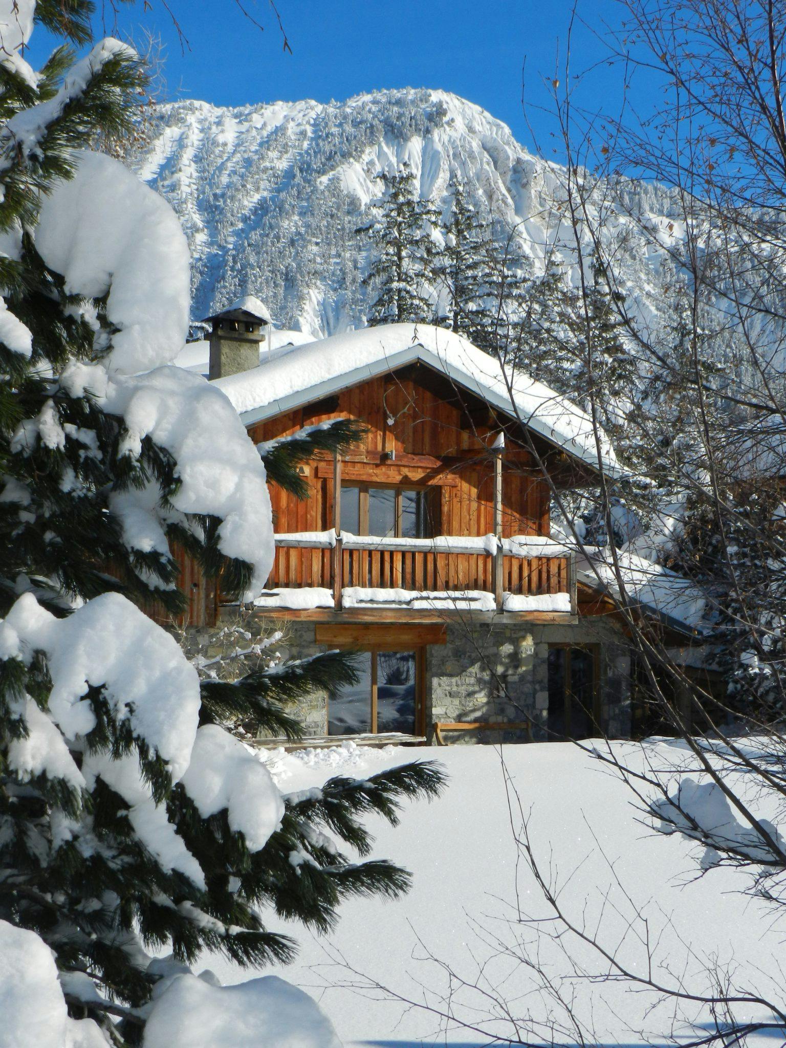 Un chalet en bois confortable avec des fondations en pierre, partiellement cachée par des arbres enneigés, est nichée dans un paysage enneigé avec une montagne imposante drapée de neige en arrière-plan. La scène est ensoleillée et sereine, évoquant une atmosphère de pays des merveilles hivernales.