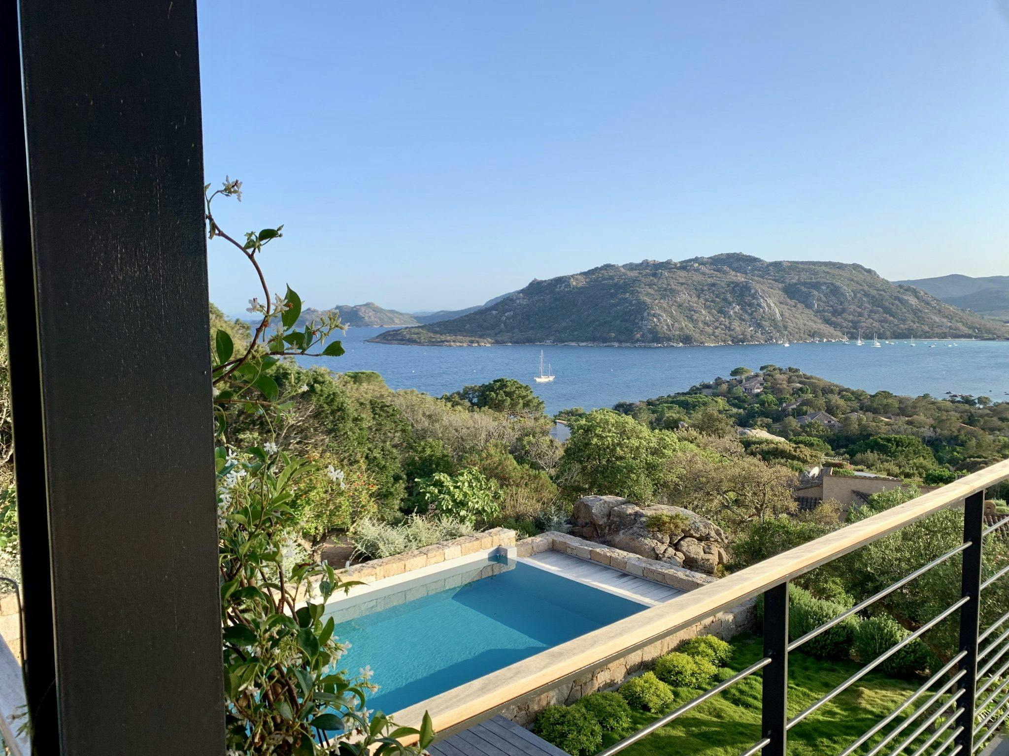 A panoramic view from a balcony overlooking an infinity pool, lush greenery, and vast blue waters in a luxury Corsican destination. In the background, hills and a few sailboats are visible under a clear, sunny sky. The balcony's railing is in the foreground.