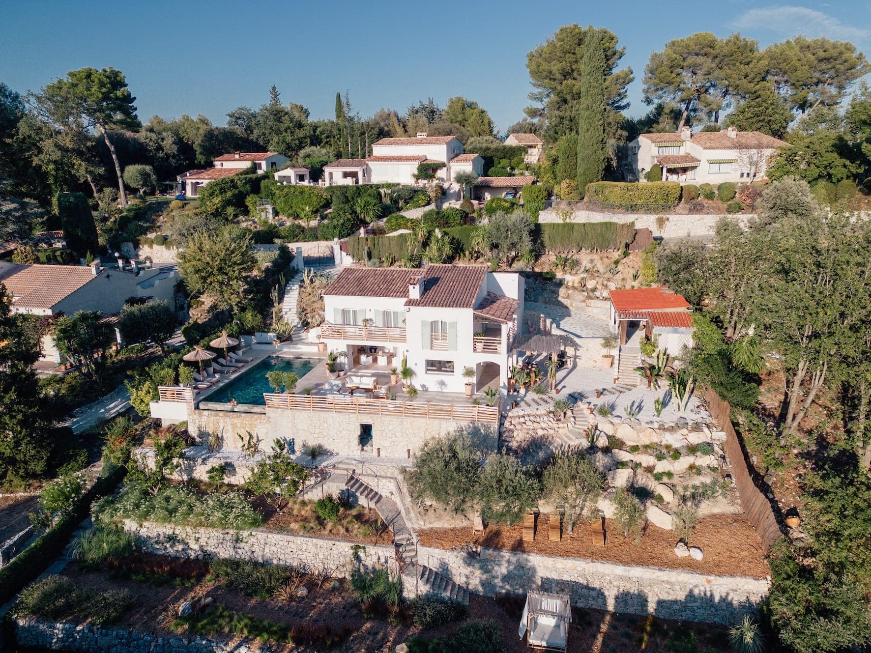 Vue aérienne d'une villa de style méditerranéen entourée de verdure et d'autres maisons. La villa dispose de jardins en terrasses, d'une piscine et de plusieurs espaces extérieurs avec vue sur un arrière-plan d'arbres et d'un ciel bleu clair.