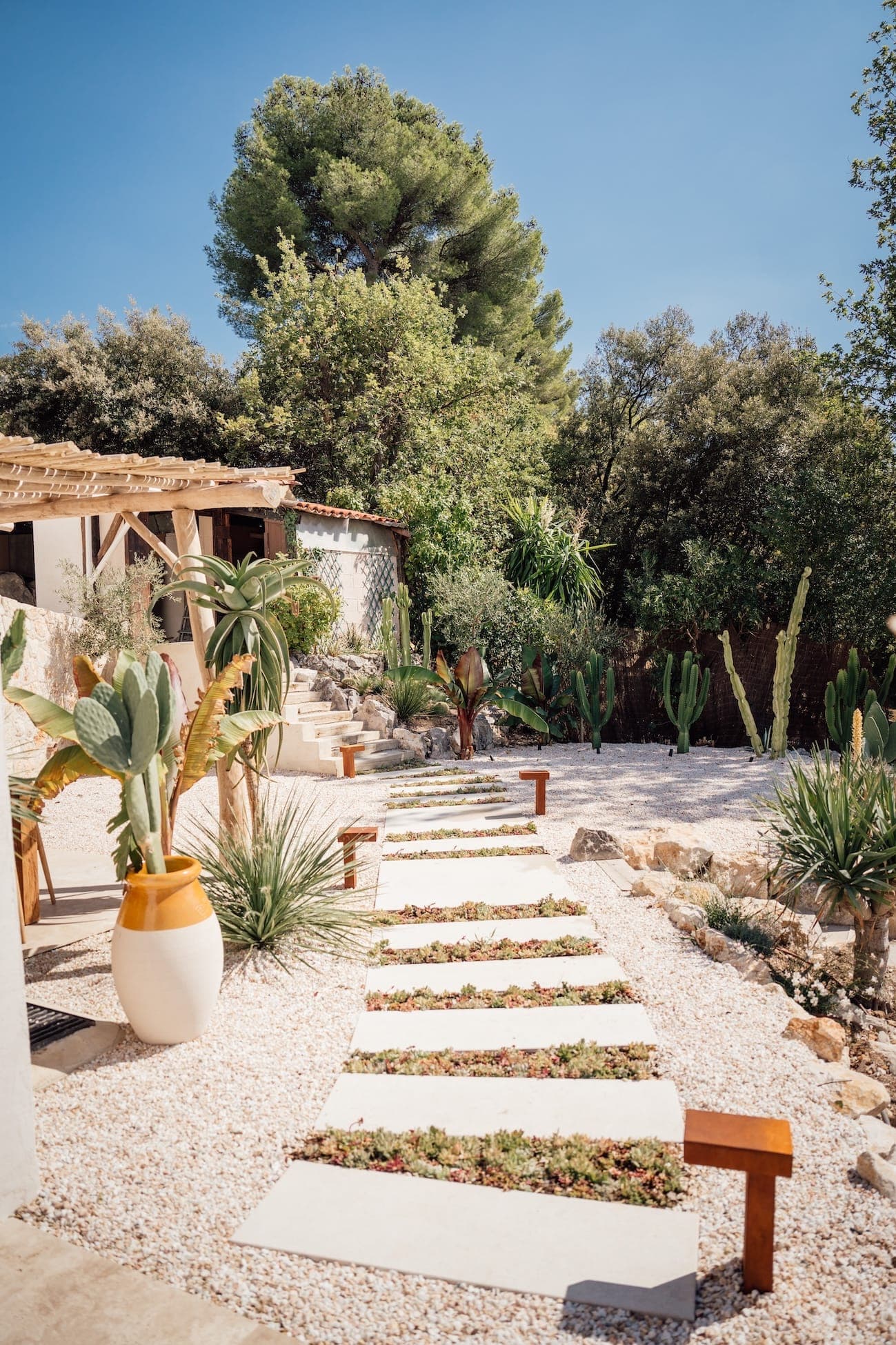 Un chemin avec de grandes dalles de pierre traverse un jardin orné de plantes succulentes, de cactus et d'autres plantes. Un bâtiment rustique avec une pergola en bois se trouve sur la gauche, entouré d'arbres et d'un ciel bleu clair au-dessus.