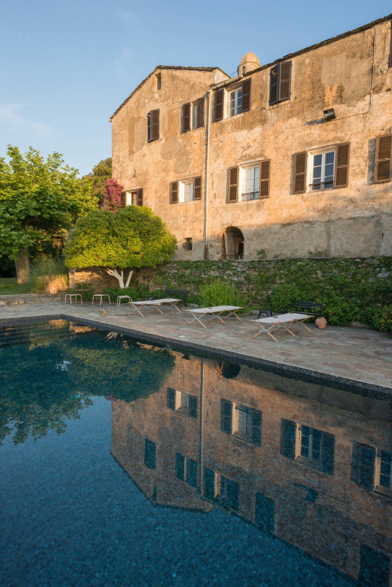 Un bâtiment en pierre aux fenêtres fermées par des volets se reflète dans une piscine bleue calme. Le bord de la piscine est doté de plusieurs chaises longues et est entouré d'arbres luxuriants et de verdure. Baigné par la lumière chaude et dorée du soleil, le paysage serein respire une ambiance rustique, ce qui en fait l'une des destinations de luxe que la Corse a à offrir.