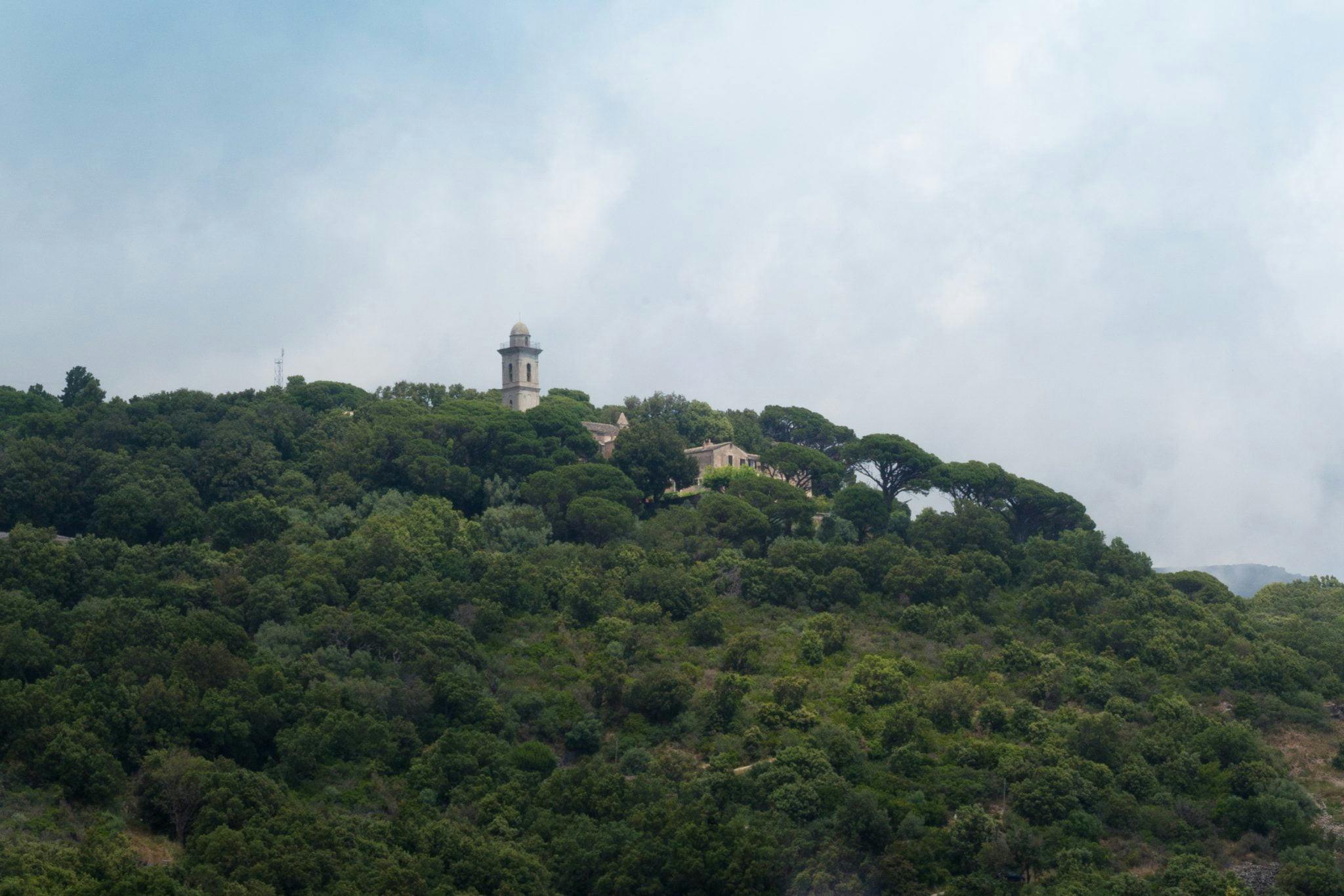 Une vue panoramique sur une colline verdoyante et luxuriante avec un bâtiment d'aspect historique doté d'une tour parmi les arbres. Le ciel au-dessus est nuageux, ajoutant un sentiment de sérénité à cette destination corse, parfaite pour ceux qui recherchent une escapade de luxe dans l'étreinte de la nature.