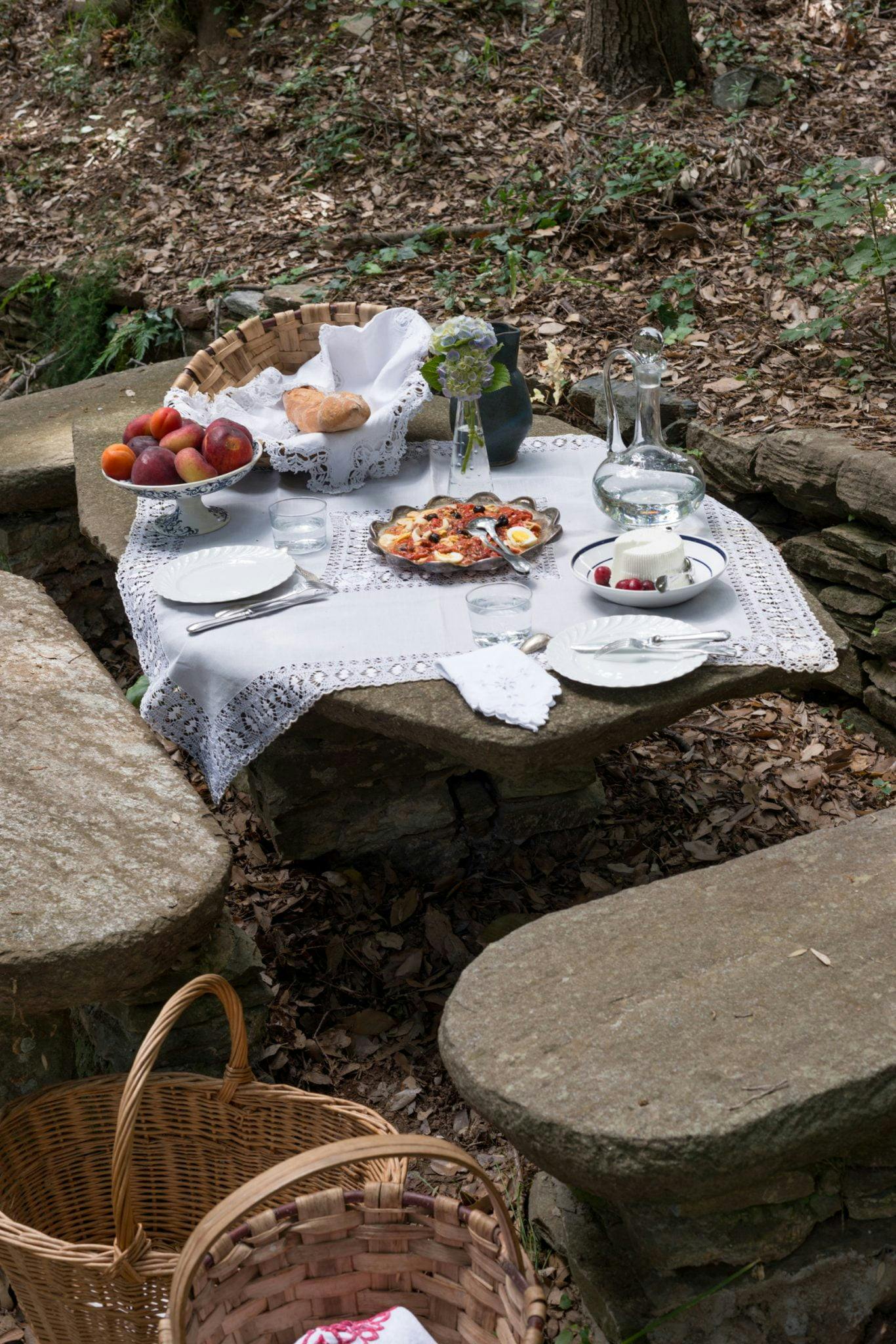 Une table de pique-nique en pierre dans une forêt, dressée avec une nappe blanche au crochet, évoque le charme des destinations de luxe. La table présente un panier de pommes, des petits pains, un pichet d'eau, des assiettes avec de la nourriture dont une tarte et un vase avec des fleurs. Autour de la table se trouvent des bancs en pierre et un panier en osier posé au sol.