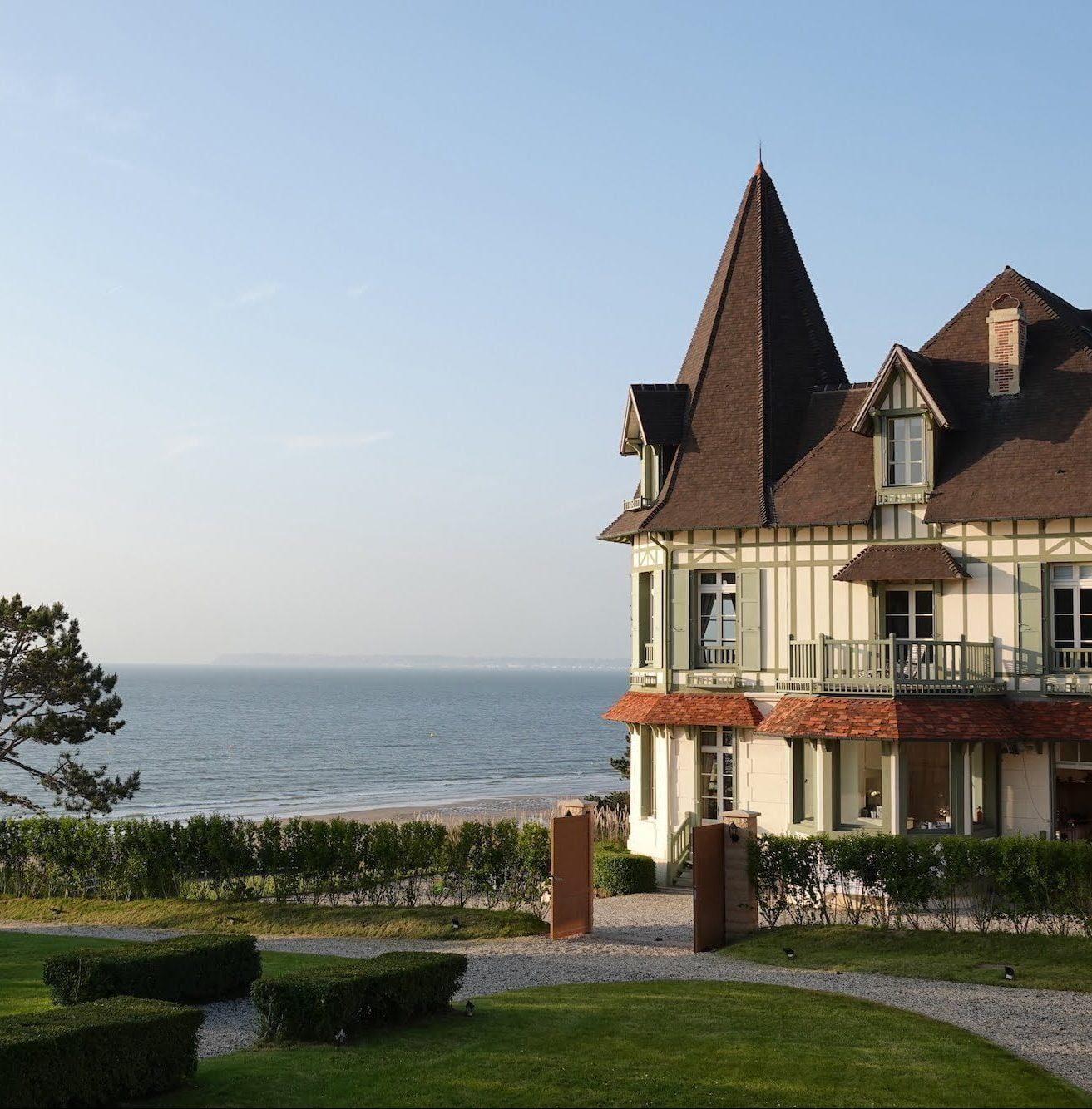 A picturesque coastal scene features a charming, large house with a pointed tower and a traditional European architectural style. The house is situated near a beach, with lush green gardens in the foreground and a calm sea in the background.