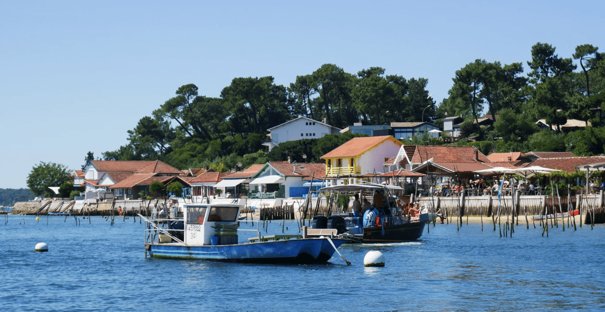 Scène de vacances sur le Bassin d'Arcachon. La scène côtière est sereine avec des bâtiments et des arbres aux couleurs vives en arrière-plan. Plusieurs bateaux flottent sur une eau bleue calme près du rivage. L'endroit parfait pour réserver une location de vacances de luxe en France.