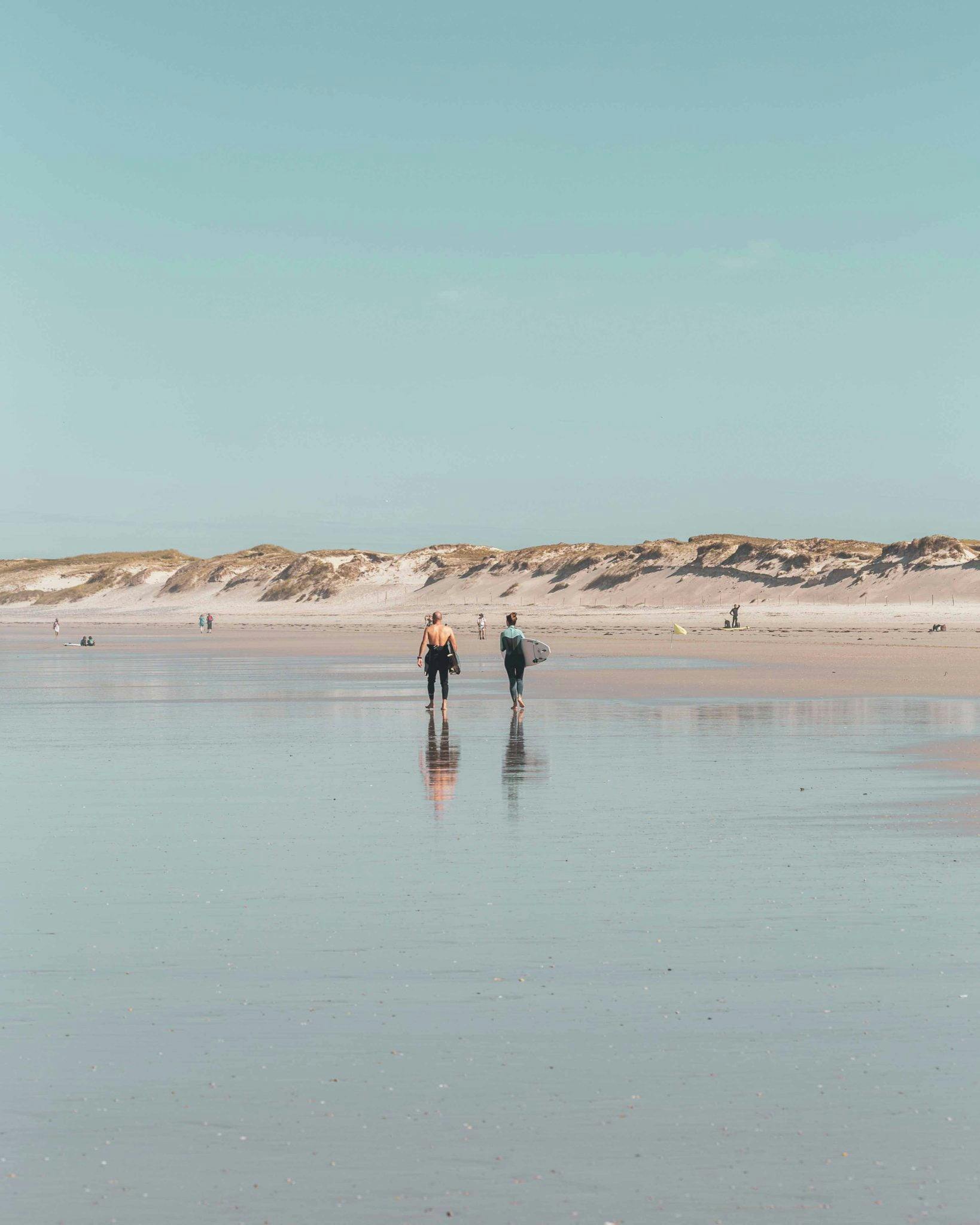 Deux surfeurs marchant sur la plage, dunes et ciel bleu en arrière plan