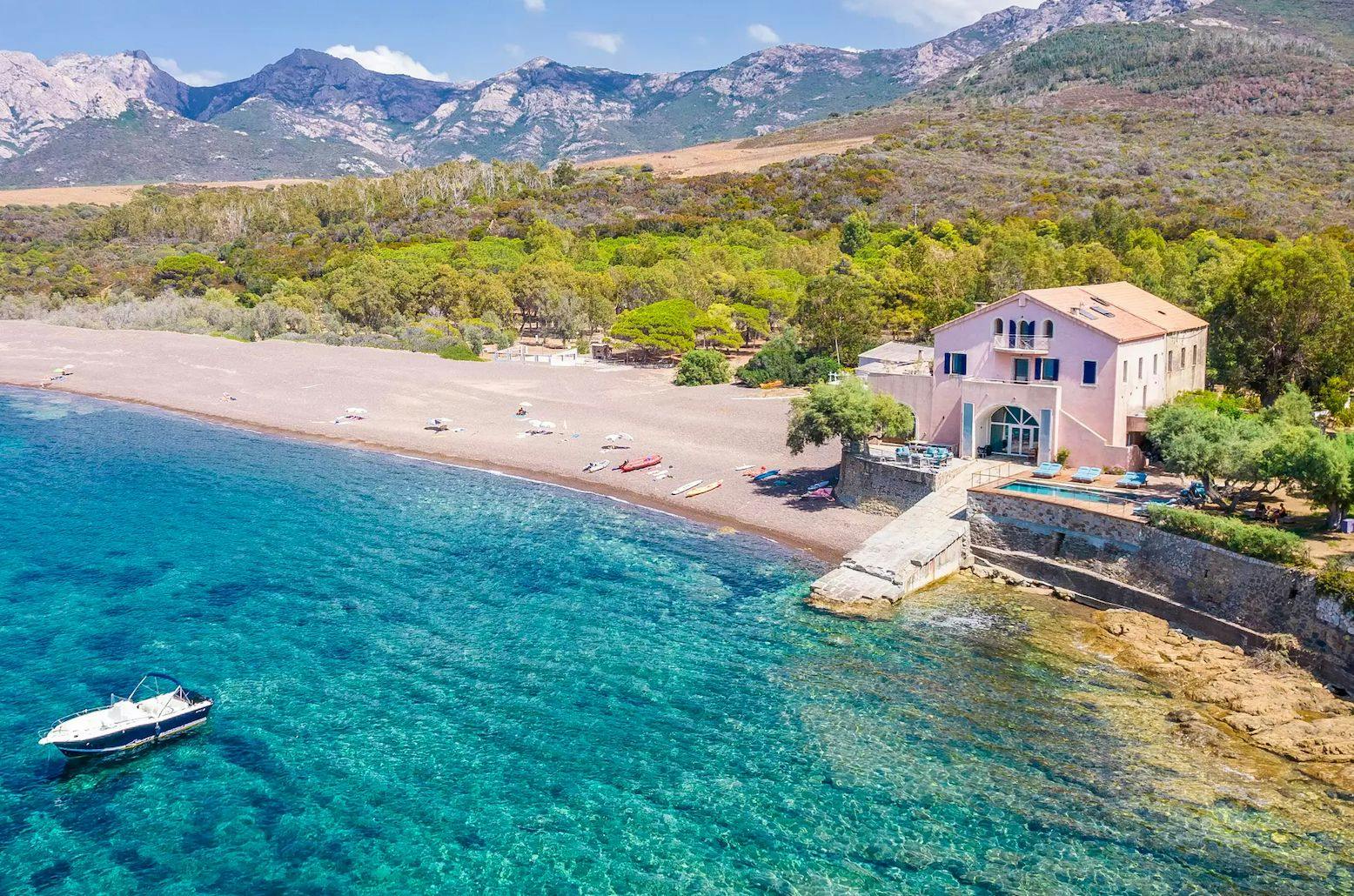 Aerial view of a tranquil coastal scene in luxurious Corsica, featuring a sandy beach bordered by lush greenery and mountains in the background. A large pink house sits on the shore next to a pier extending into the clear, turquoise water where a white boat is anchored. An idyllic destination awaits.