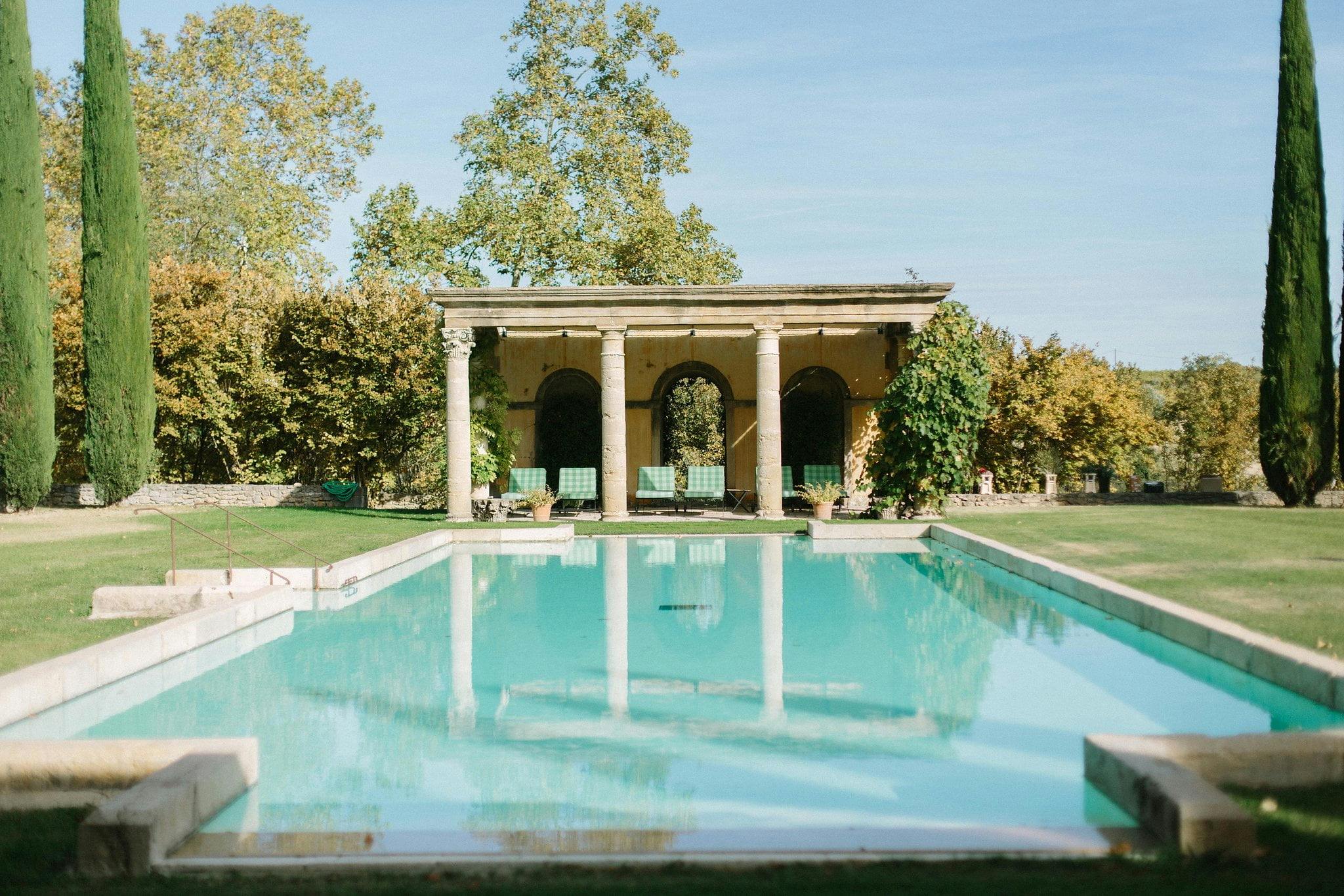 The serene rectangular swimming pool of Château de Sannes - one of the best luxury vacation rentals in France - is flanked by green lawns and tall cypress trees. At the far end, a stone pavilion with three arches stands, providing a shaded seating area. The clear blue sky and lush greenery are reflected in the calm pool water.