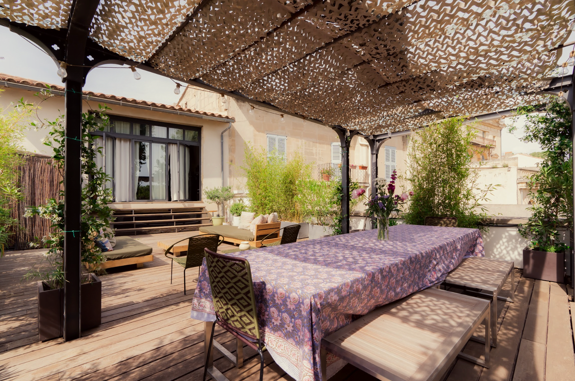 A cozy outdoor patio with a shaded pergola, featuring a long wooden dining table covered with a purple-patterned tablecloth. A bench and several chairs surround the table. In the background, there are cushioned seating areas, plants, and a view of a house with large windows.