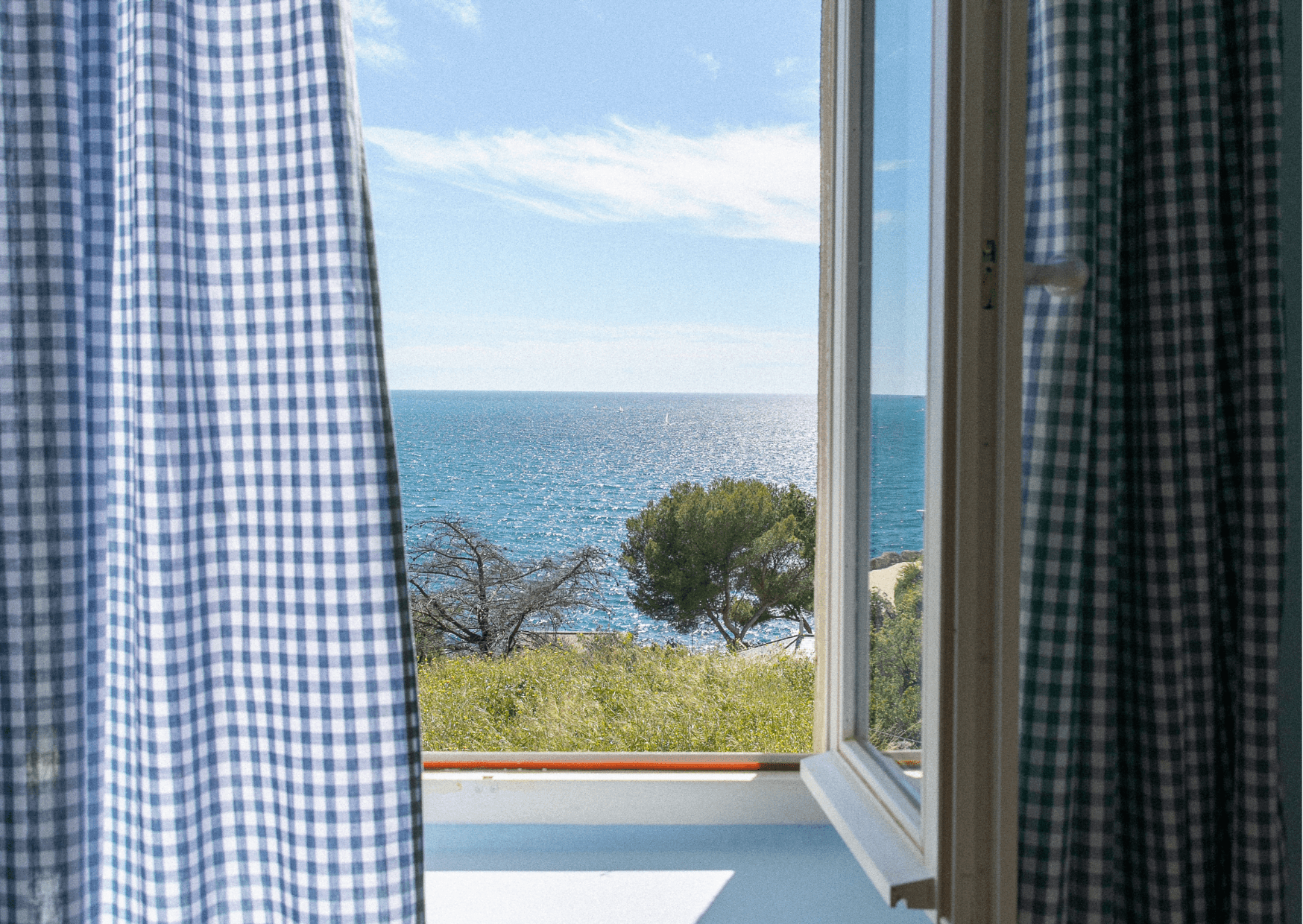 Admire the serene view from a villa in Marseille through an open window adorned with blue and white checkered curtains. Admire the picturesque ocean landscape, framed by trees and grassy fields under a clear blue sky.