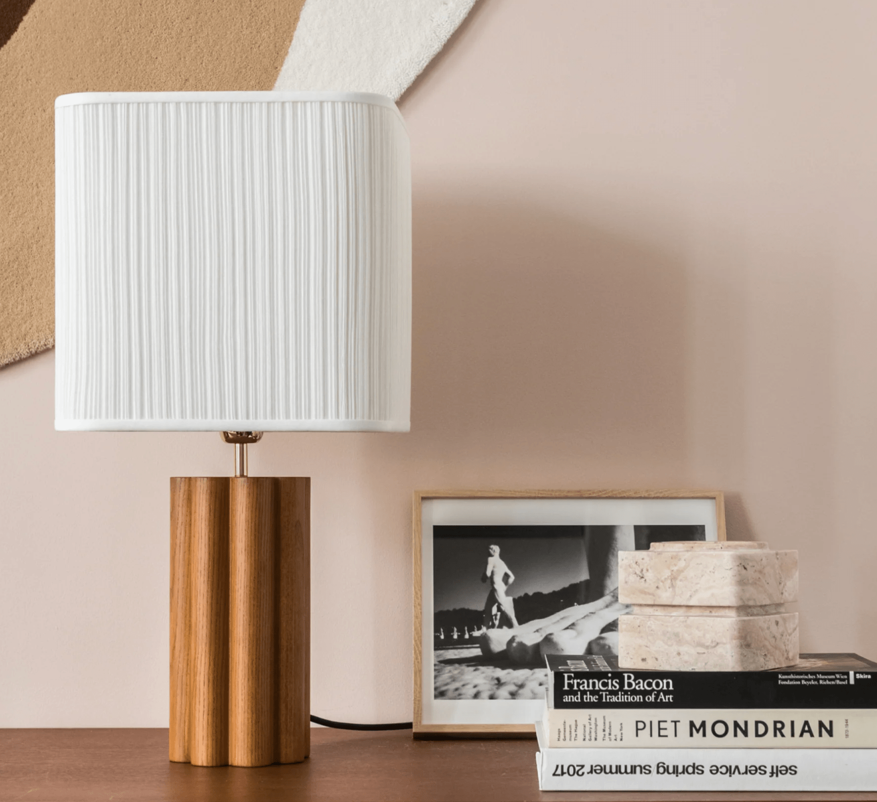 Une lampe de table moderne avec une base en bois et un abat-jour plissé blanc se trouve sur une surface en bois à côté d'une pile de livres et de boîtes rondes blanches. Une photo encadrée en noir et blanc est placée derrière les objets contre un mur de couleur claire orné d'art abstrait.