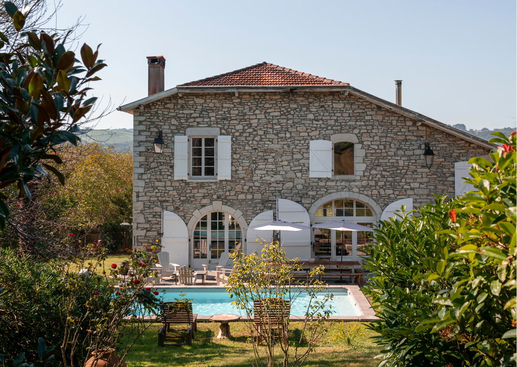 A charming stone house with white shutters stands next to a swimming pool surrounded by a garden. Two lounge chairs face the pool, and a tall umbrella provides shade. Trees and plants frame the scene, and the landscape in the background completes the idyllic setting.