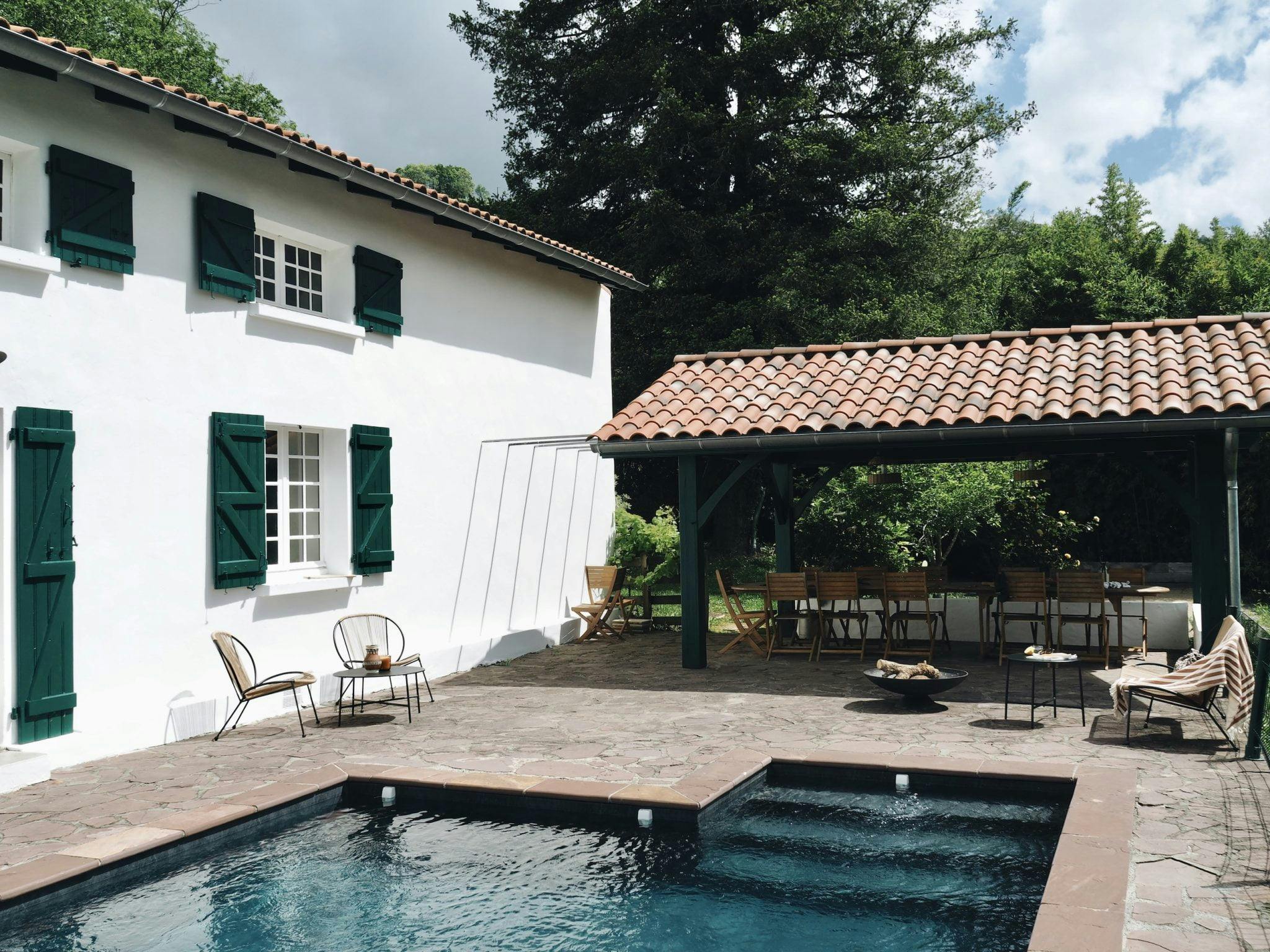 Une cour sereine avec une piscine rectangulaire bordée d'un patio en pierre. A côté de la piscine se trouve une maison blanche aux volets verts. À droite, un belvédère ombragé au toit de tuiles abrite un coin repas avec des chaises et une table en bois, entouré d'une verdure luxuriante.