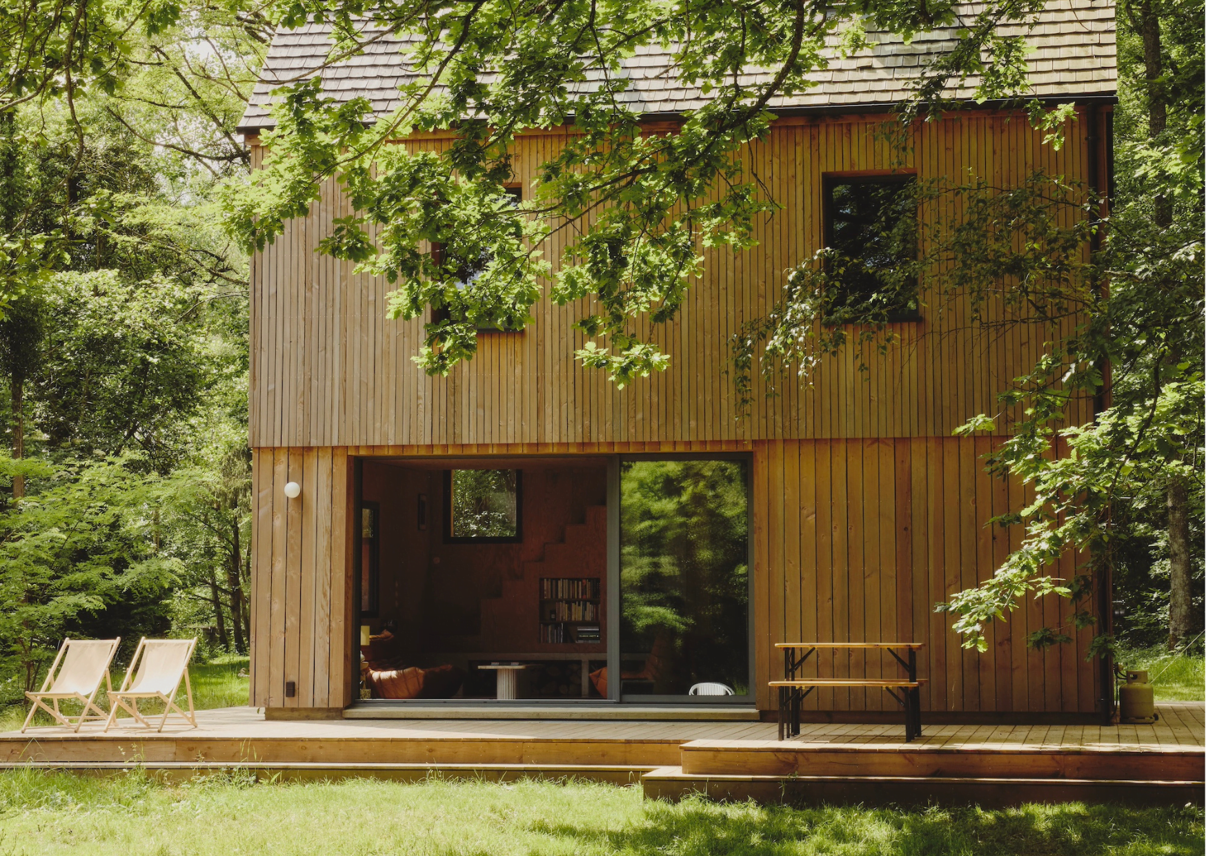 Une maison en bois à deux étages avec de grandes fenêtres nichée dans une forêt luxuriante. Il y a une grande terrasse en bois avec deux transats et un banc. De grands arbres verts entourent la maison et la lumière du soleil filtre à travers les feuilles, créant une atmosphère sereine.