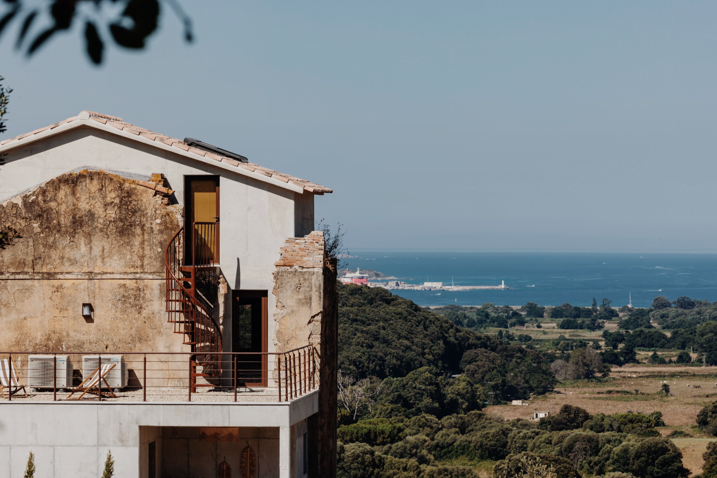 The facade of the renovated house, with an iron staircase leading up to the terrace on the upper floor. Fields, forests, and the sea in the background.