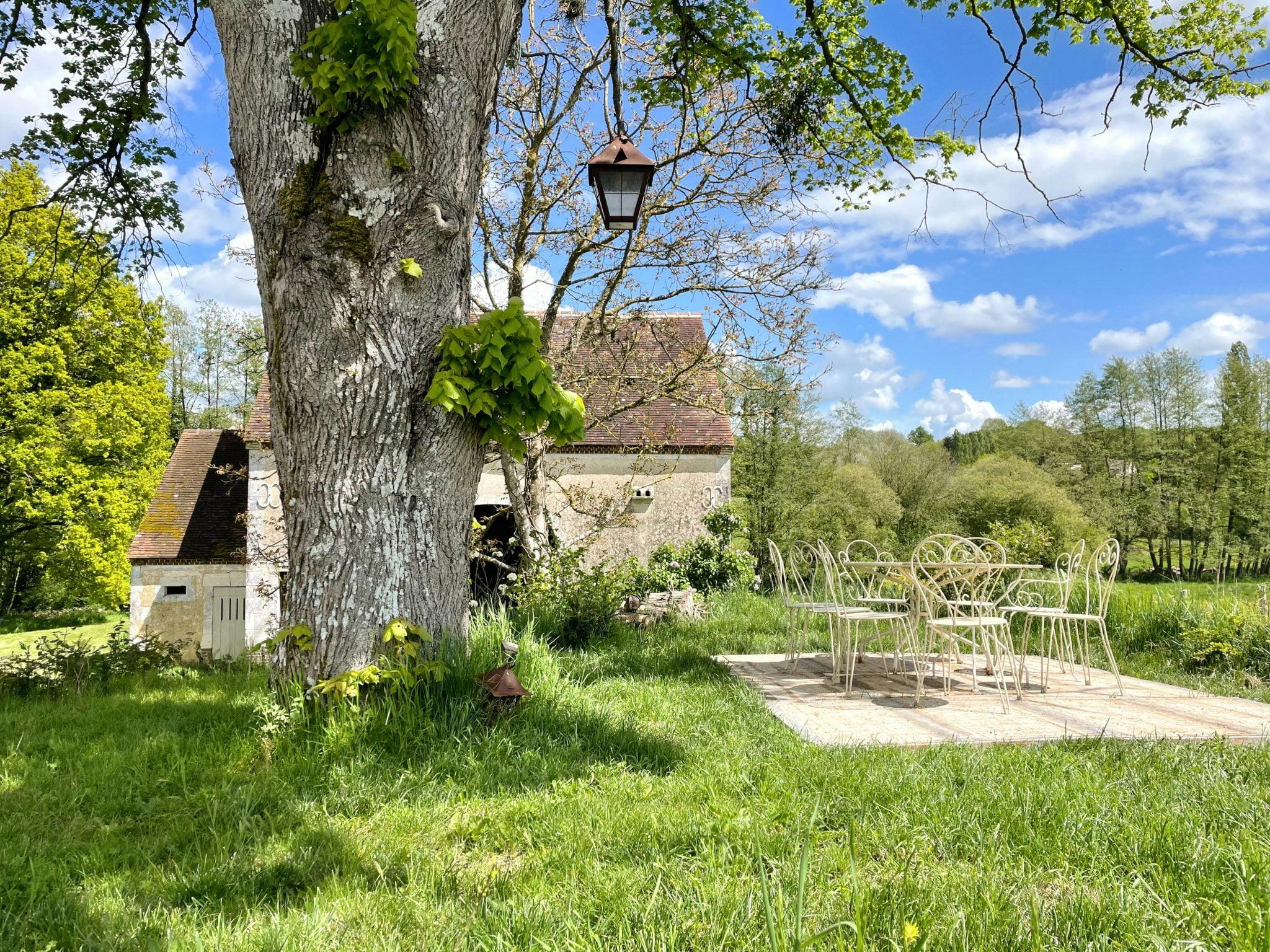 Une scène extérieure paisible montre un patio avec une table et des chaises en métal vintage placées sous un arbre aux nouvelles feuilles printanières. Un bâtiment rustique en pierre avec un toit abrupt se trouve à l'arrière-plan et les environs sont luxuriants et verdoyants avec des arbres et de l'herbe, baignés de soleil.