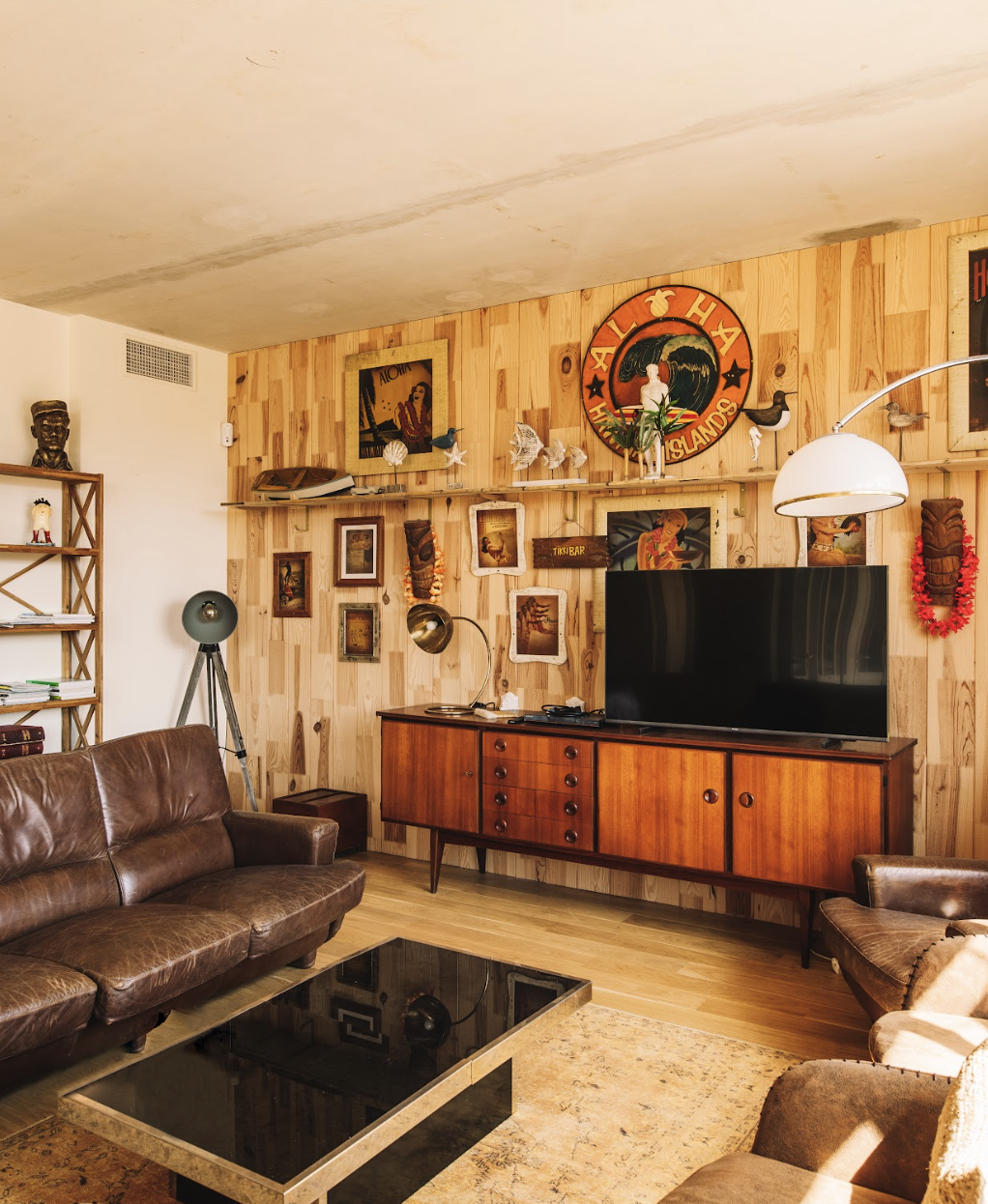 A cozy living room with wooden walls adorned with various vintage decorations, such as framed paintings, masks, and a Hawaiian sign. The room features a large brown leather sofa, a black coffee table, a flat-screen TV on a wooden console, and a floor lamp.