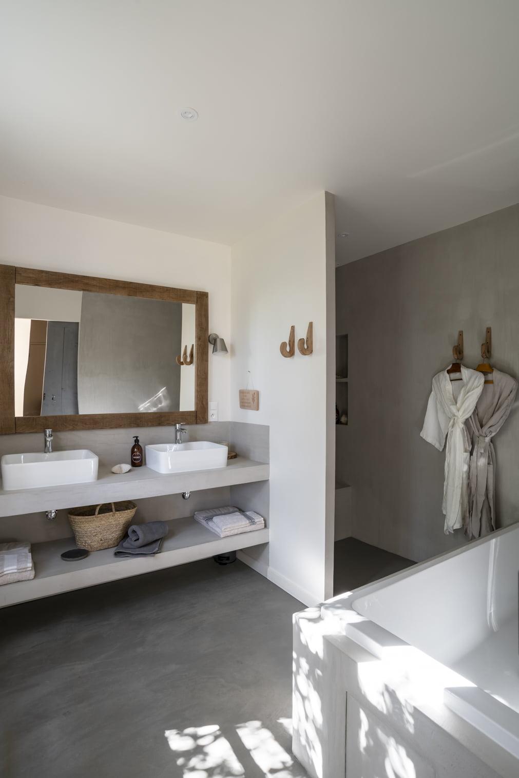 Une salle de bains moderne au design minimaliste comprend un grand miroir avec cadre en bois au-dessus de deux lavabos blancs. Sous les éviers se trouvent des étagères contenant des serviettes et un panier en osier. Des peignoirs sont suspendus à des crochets en bois à côté d'une douche fermée et une baignoire est au premier plan.