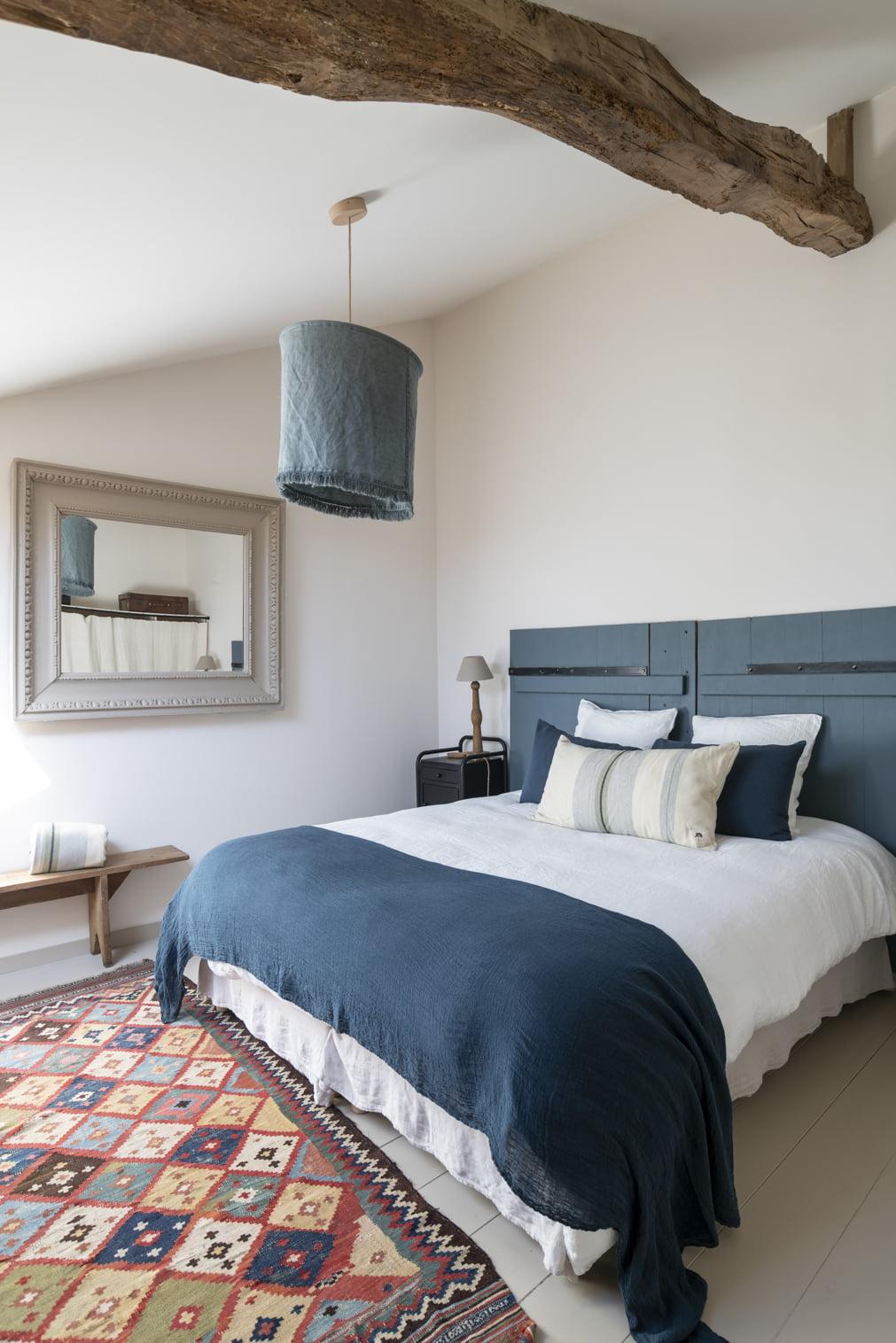 A cozy bedroom featuring a bed with blue and white bedding, a large wooden beam on the ceiling, and a textured blue lampshade. A patterned rug adds color, and a large mirror and a wooden bench enhance the decor. A small lamp sits on a bedside table.