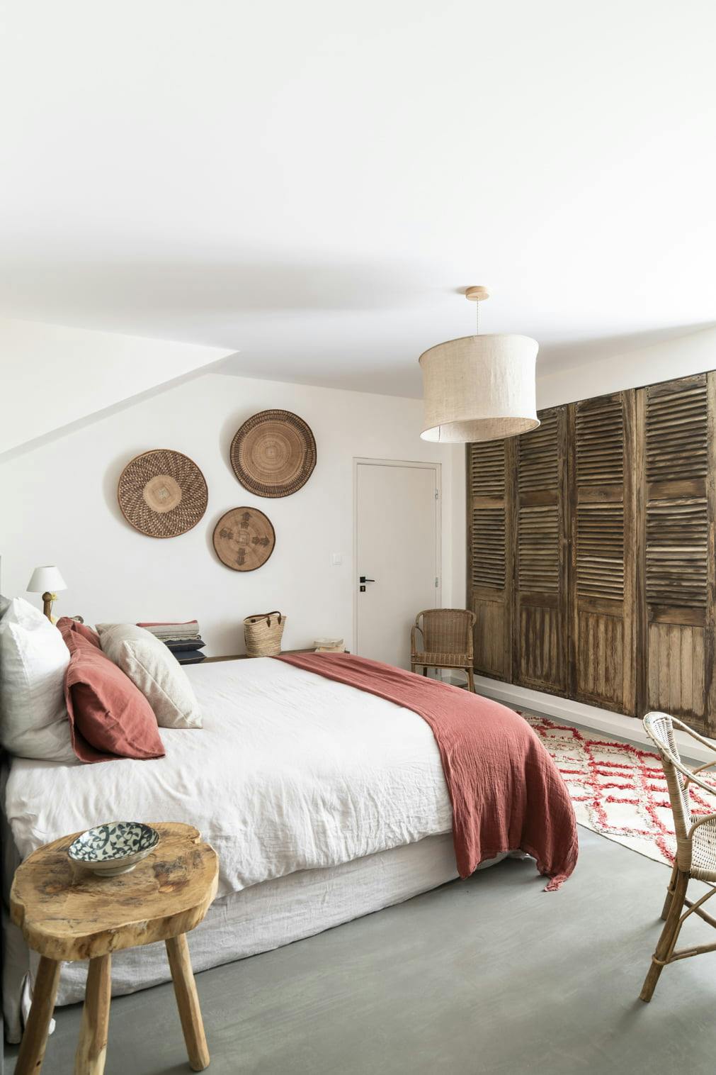 A cozy bedroom with a bed dressed in white bedding and a red throw. Three wooden wall decorations are hung above the bed. The room features a rustic wooden sliding closet door, a small wooden side table, a woven chair, and a soft beige light fixture.