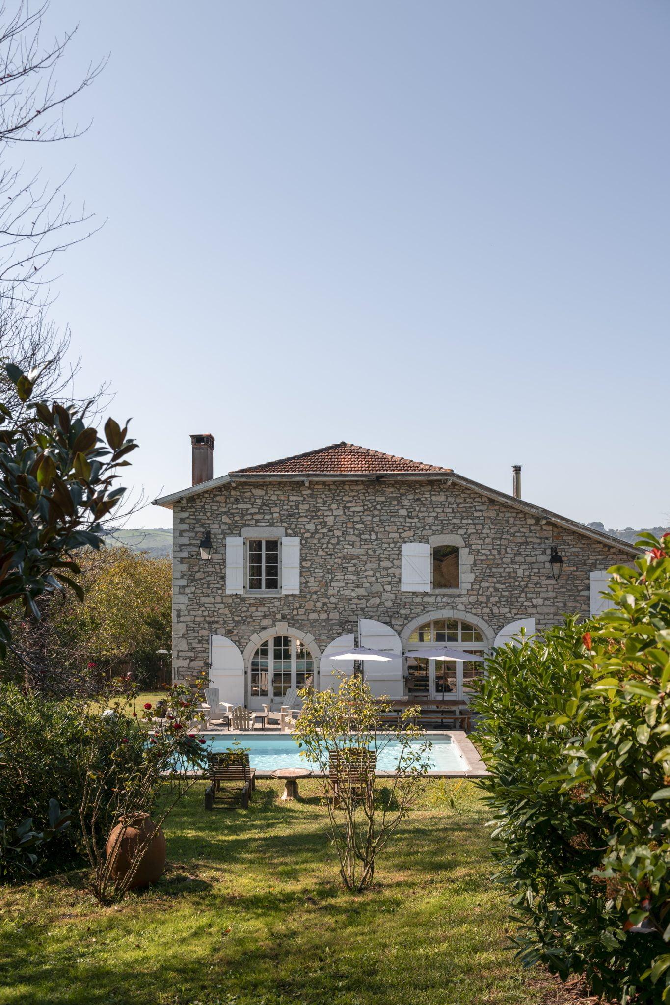 A charming stone house with a red tiled roof, featuring arched windows and white shutters. In front of the house is a pool surrounded by a lush garden with various plants and trees under a clear blue sky.