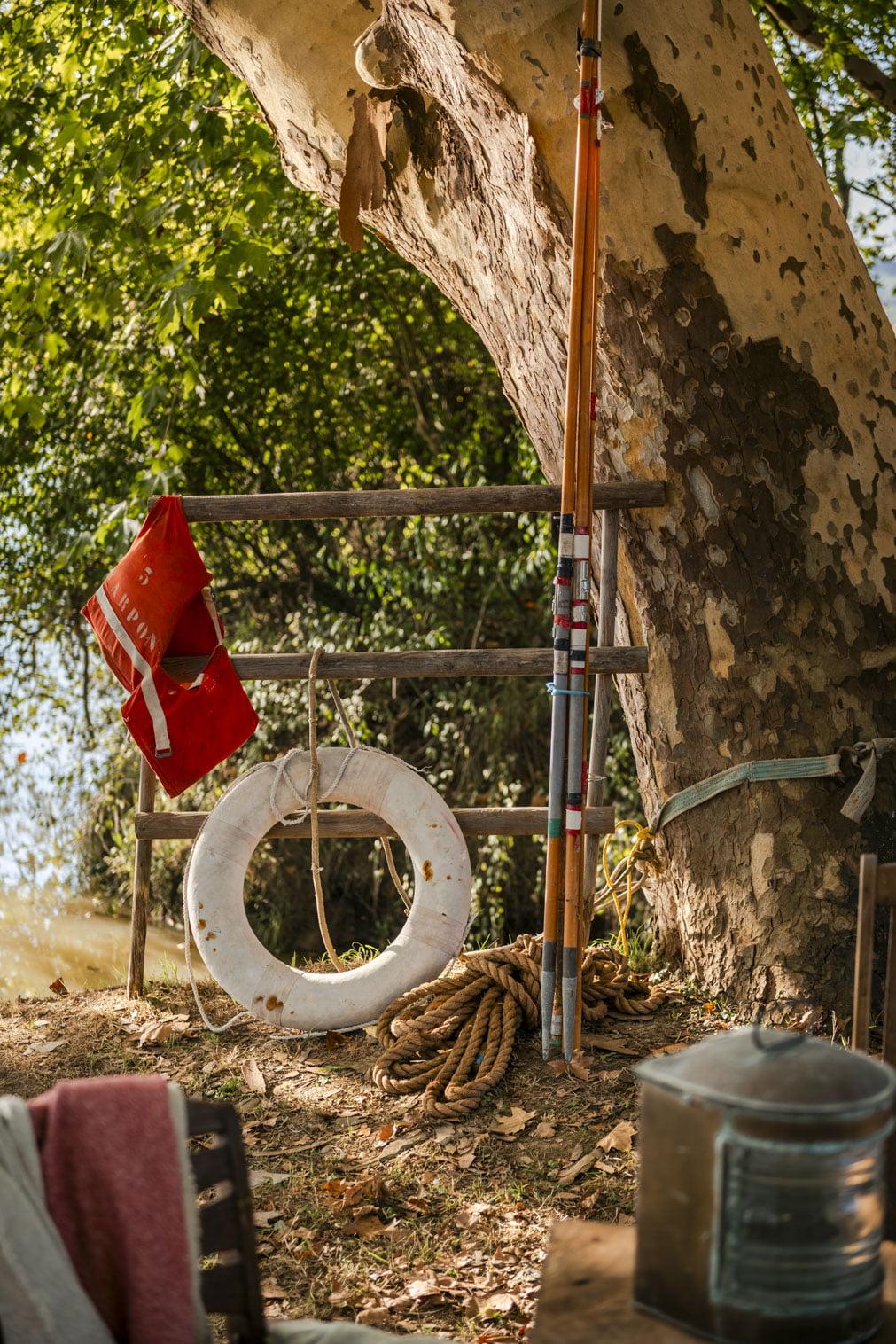 A weathered life buoy and an orange life jacket are hanging on a wooden fence under a tree, next to a coiled rope and two fishing rods. The area is shaded by the leaves of the trees, with a glimpse of water in the background. An old lantern is nearby, suggesting a rustic outdoor decor.