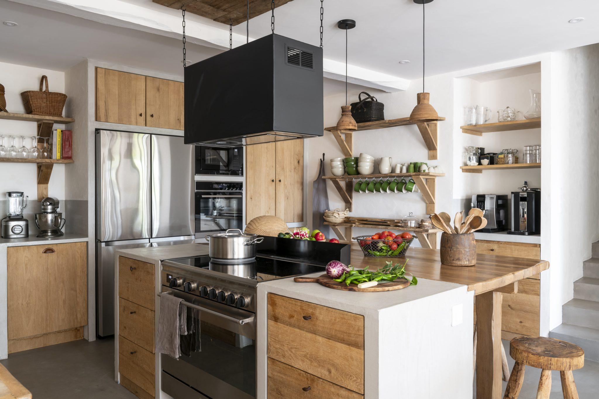 A modern kitchen with wooden cabinets, stainless steel appliances, and open shelves displaying various dishes and decorations. There is an island with two bowls of vegetables and a cutting board. A black range hood hangs above the stove, and cooking utensils are organized on the counter.