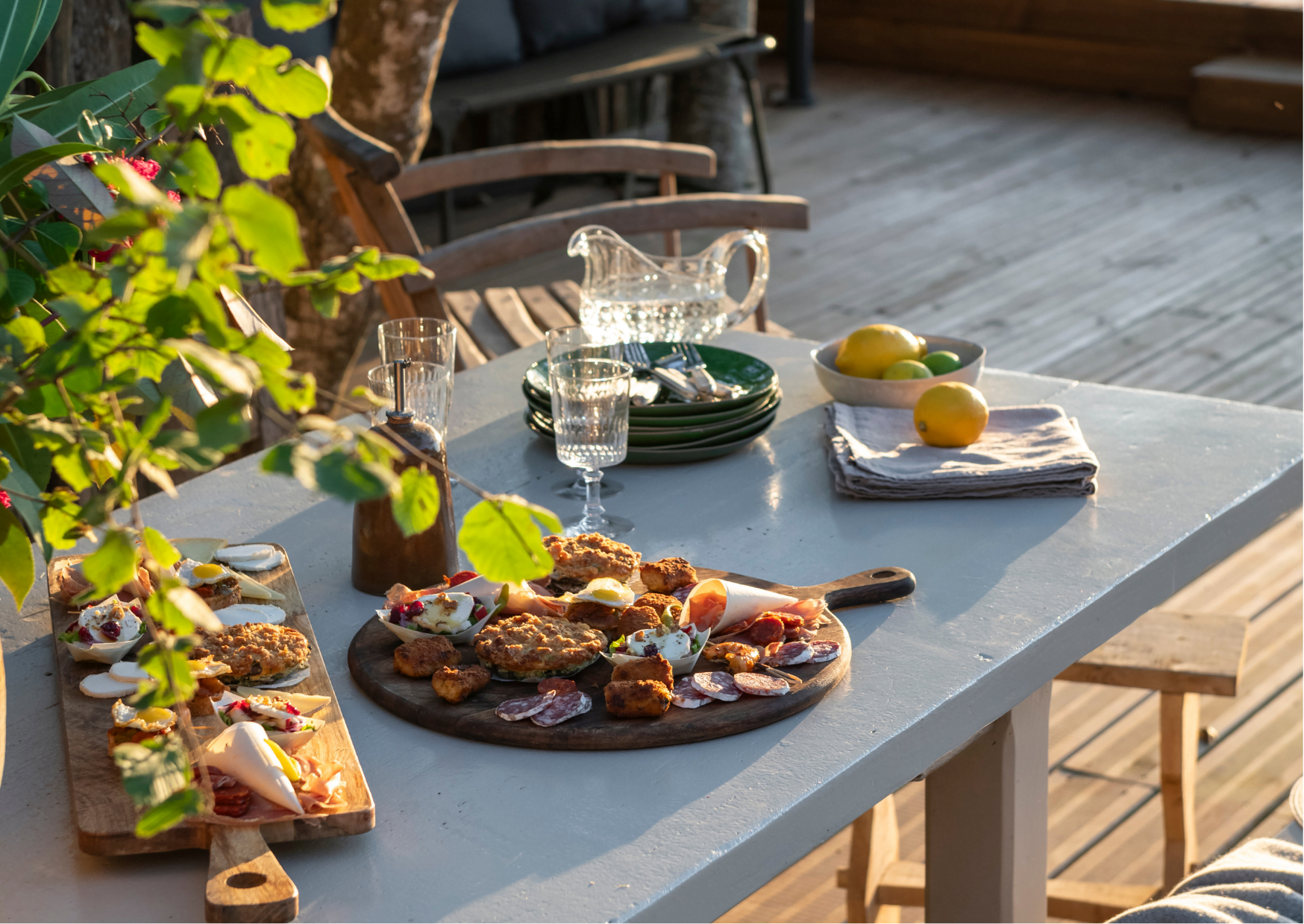 A sunny outdoor dining table for a meal features a variety of appetizers on wooden boards, stacked green plates next to a glass water pitcher, glasses filled with water, a bowl of lemons, and folded napkins. The table is surrounded by chairs and a rustic wooden deck.