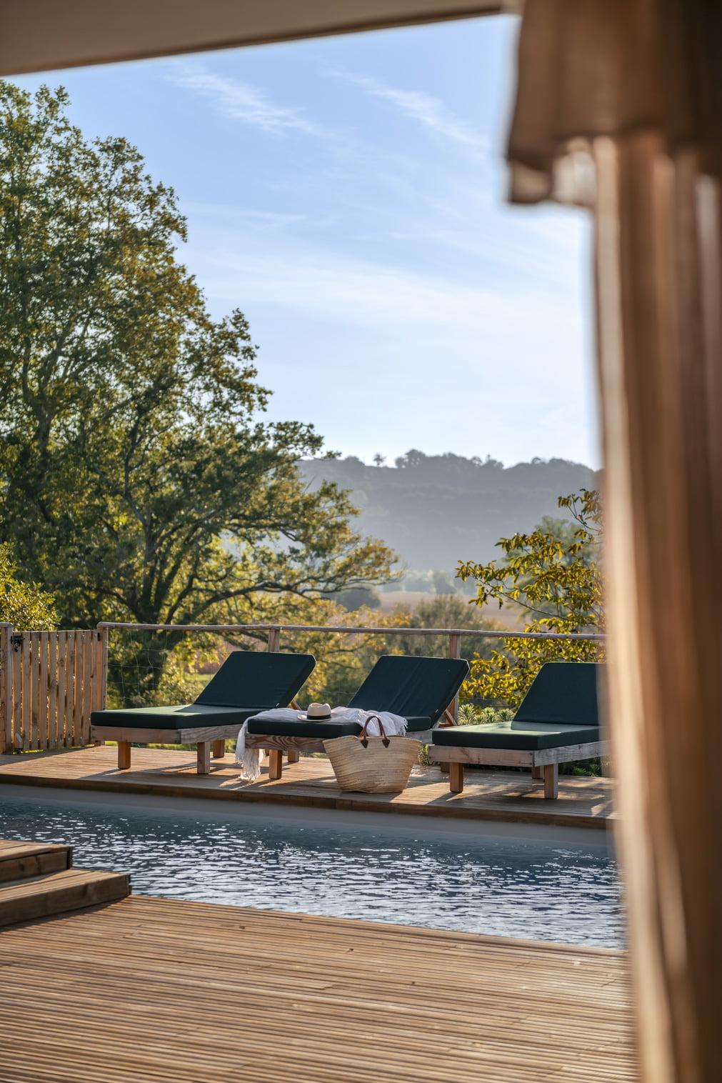 Une piscine extérieure sereine avec trois chaises longues vides face à un paysage boisé et des collines lointaines. Un panier tressé avec des serviettes et des tongs se trouve sur la terrasse de la piscine. La scène est encadrée par des rideaux sur les bords, suggérant un endroit confortable et ombragé.