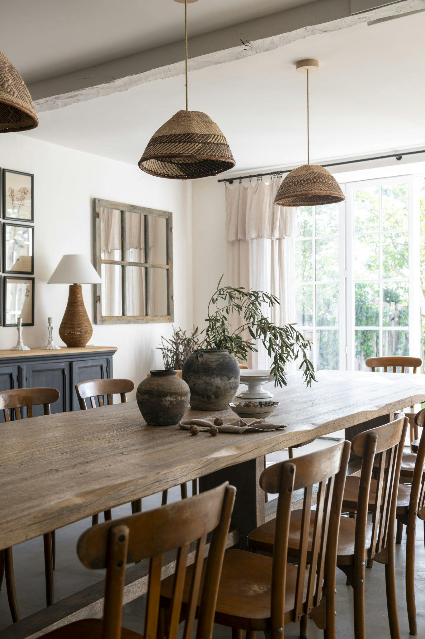 A dining room featuring a long wooden table surrounded by wooden chairs. The table is adorned with rustic clay pots and green foliage. Natural light streams in through large windows with sheer curtains. The room also showcases wicker pendant lights and framed artwork.