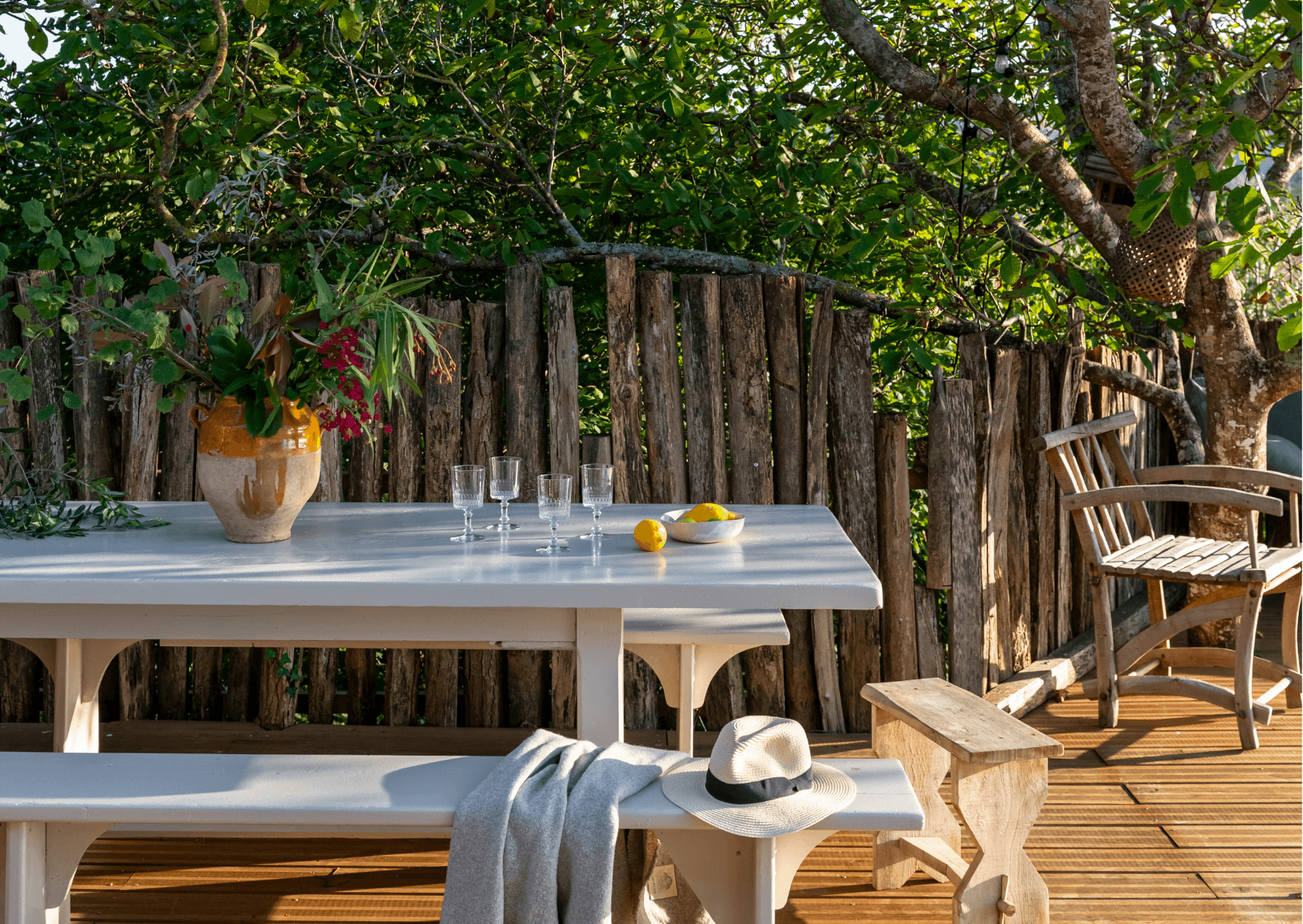 A serene outdoor setting with a wooden table and benches. The table holds a vase of flowers, some glasses, and lemons. A hat and a light garment rest on the bench. In the background, there is a rustic wooden fence and a chair, surrounded by lush greenery.