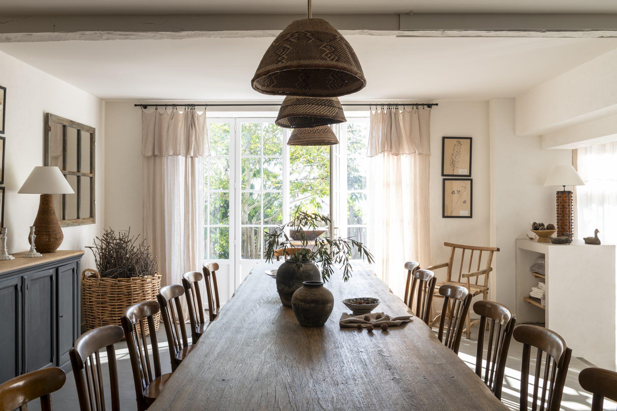 A bright dining room with a long wooden table surrounded by brown chairs. Several woven pendant lights hang from the ceiling. The room features large windows with sheer curtains, allowing natural light to flood in. The decor includes pottery, plants, and rustic accents.