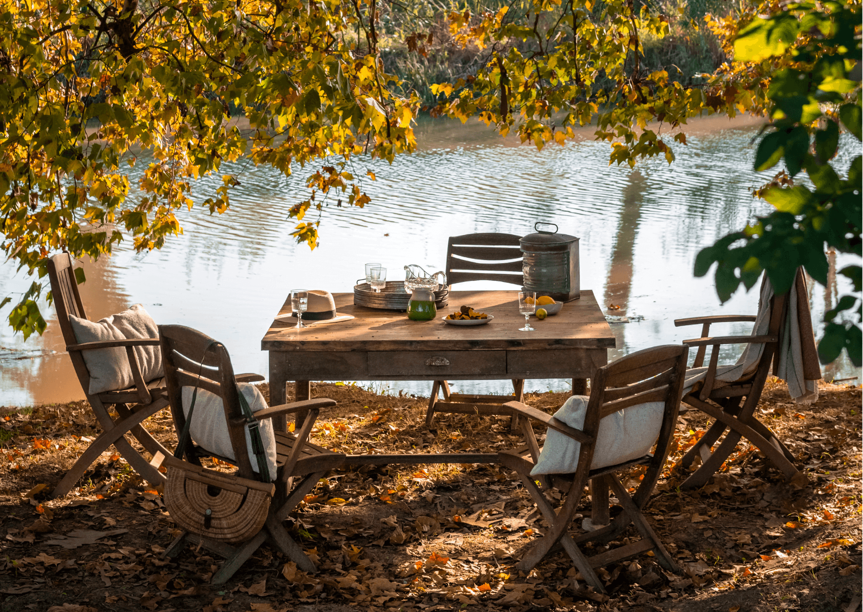 An outdoor dining table made of rustic wood located by a river in autumn. The table is adorned with glass pitchers, a vintage lantern, and plates of food. Four wooden chairs with cushions surround the table, and golden leaves hang above, casting a warm glow.