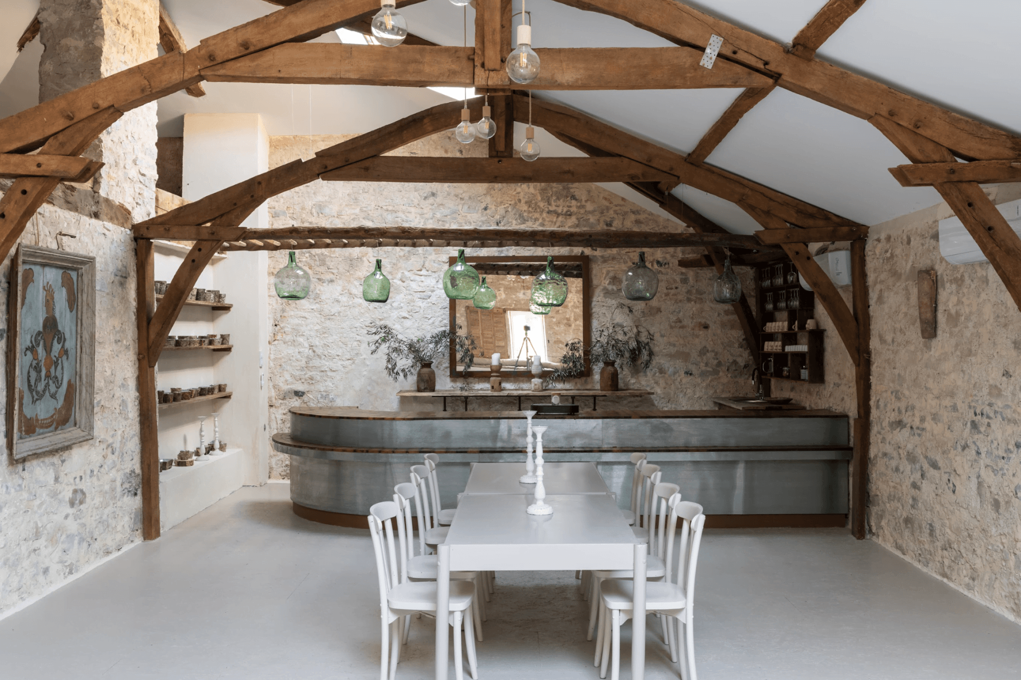 A rustic kitchen with exposed beams and stone walls features a long dining table with white chairs. The kitchen counter is sleek and modern, lined with pendant lights. Wooden shelves display various objects, and a large rectangular mirror is hung above the counter.