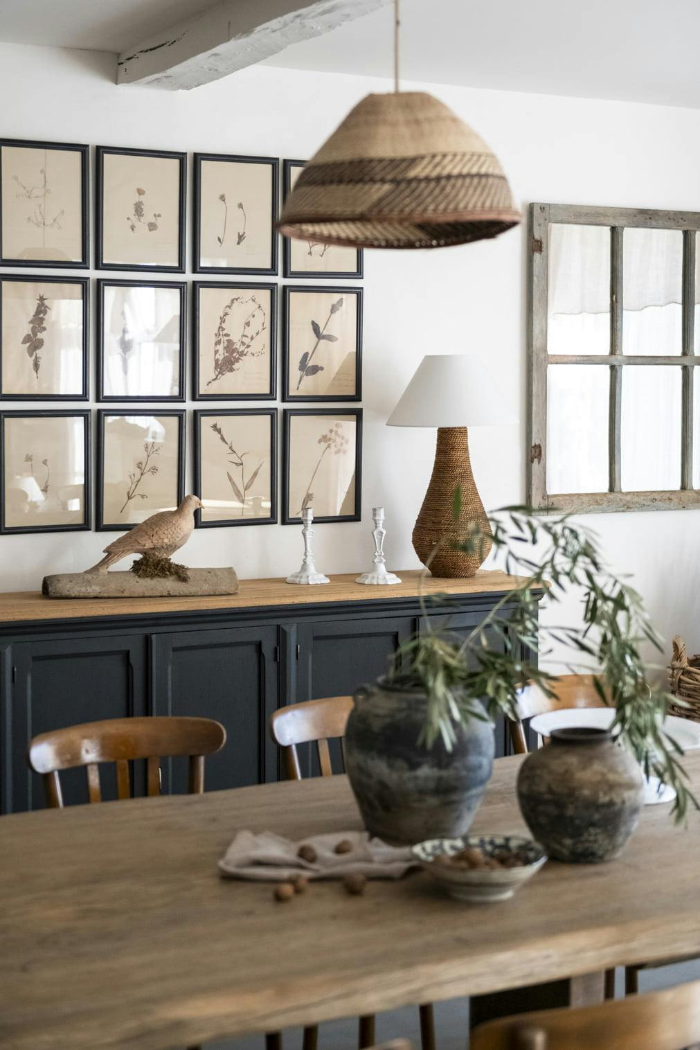 A cozy dining room featuring a wooden table with ceramic vases and a cloth on top. Behind the table is a dark-colored cabinet adorned with botanical engravings and small sculptures. A wicker pendant light hangs above, and a decorative lamp rests on the cabinet.