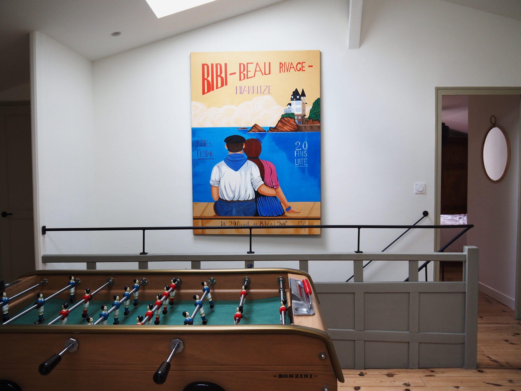 A foosball table with red and blue players is situated in a bright and open loft. Above it hangs a colorful poster depicting an illustrated couple facing each other, with text in French. The room features light wood flooring and white walls, with a door on the right side.