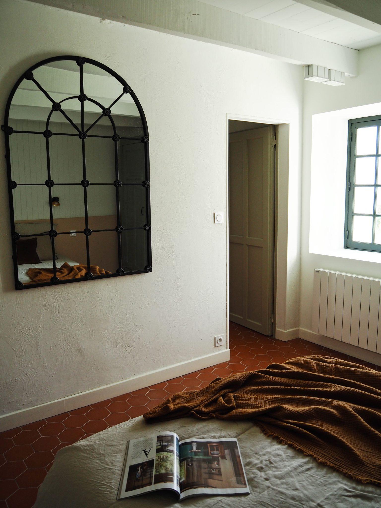 A cozy bedroom with terracotta flooring and white walls features an arched black-framed mirror hung above a bed covered with a white blanket. An open magazine and a brown throw blanket are placed on the bed. A black-framed window allows natural light to flood in.