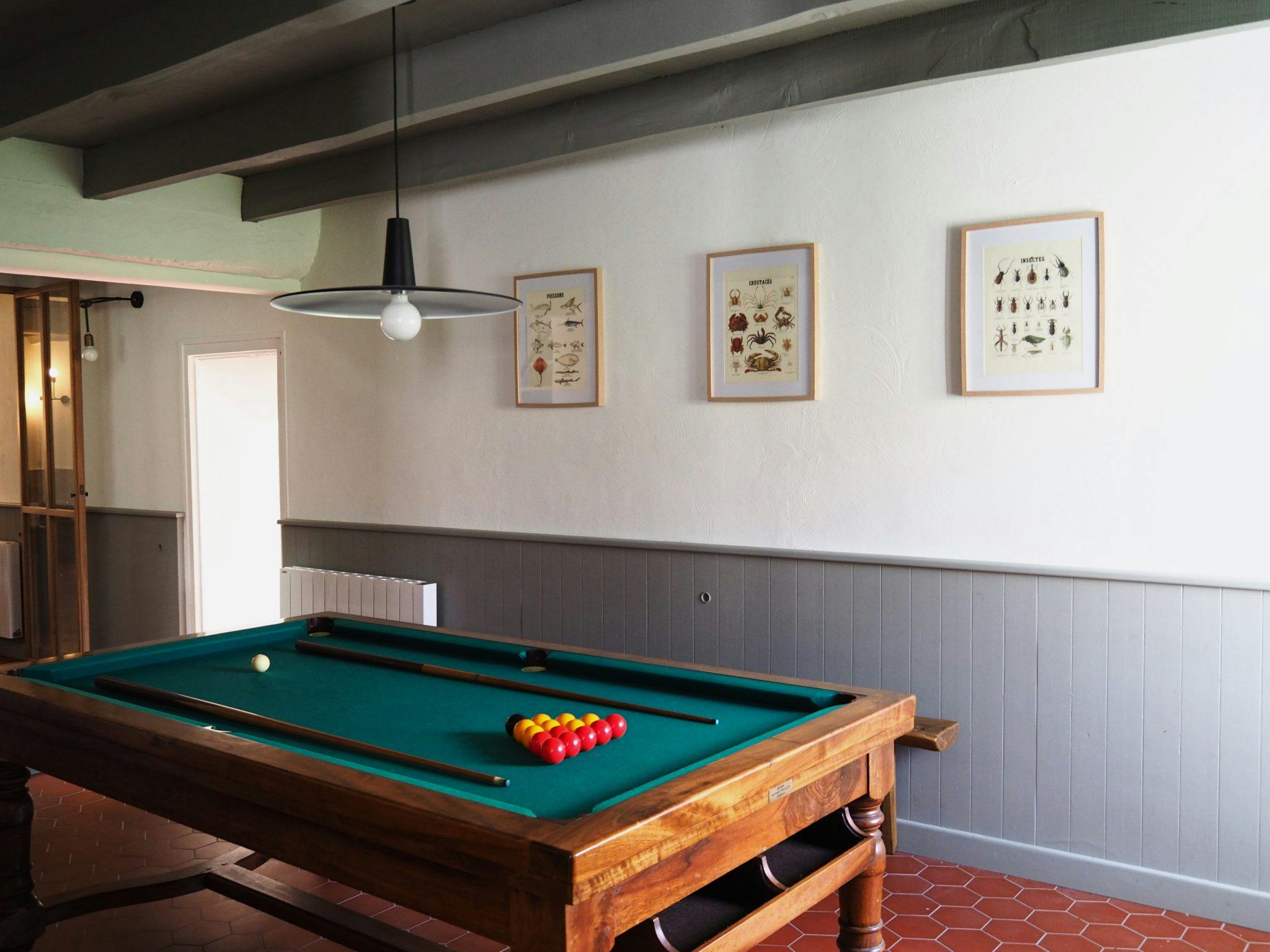 A small, well-lit room with a billiard table at the center. The billiard table has a green felt surface and the walls are decorated with framed artwork. A hanging light fixture is above the table. The room features a partially open door and a tiled floor.