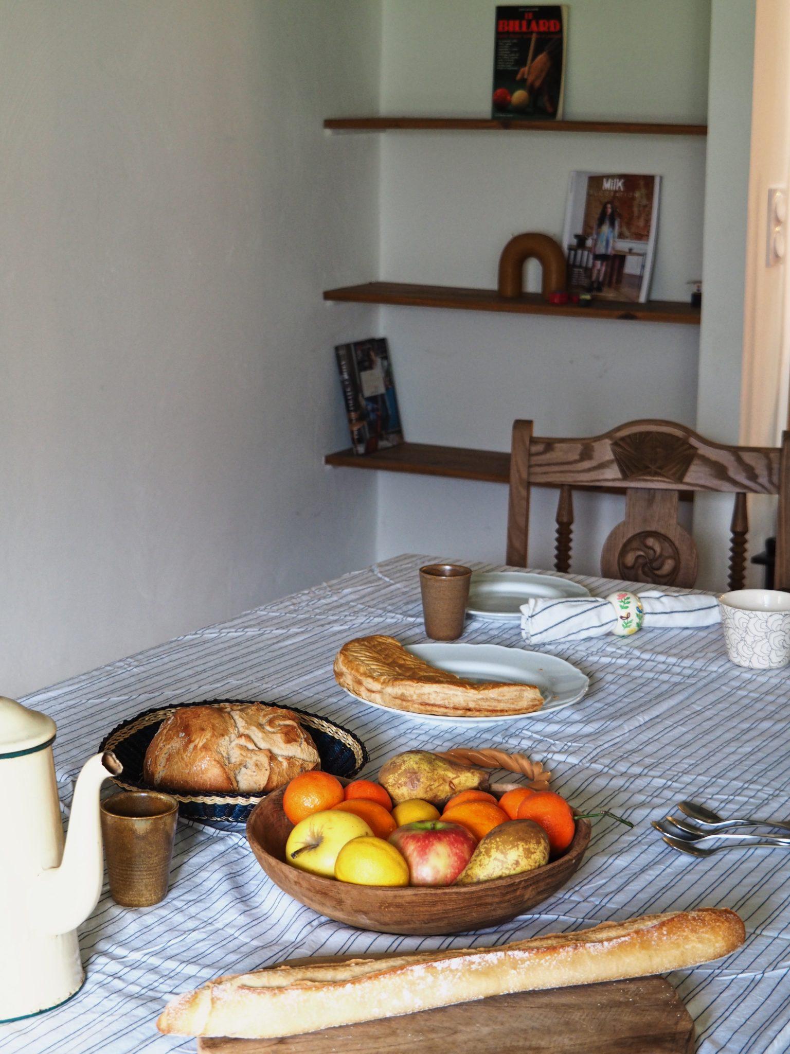 Une table à manger avec une nappe à carreaux est dressée avec une variété de plats pour le petit-déjeuner, notamment une miche de pain, une baguette, un bol de fruits mélangés, un croissant dans une assiette et une cafetière avec des tasses. Au fond, des étagères en bois exposent des livres.