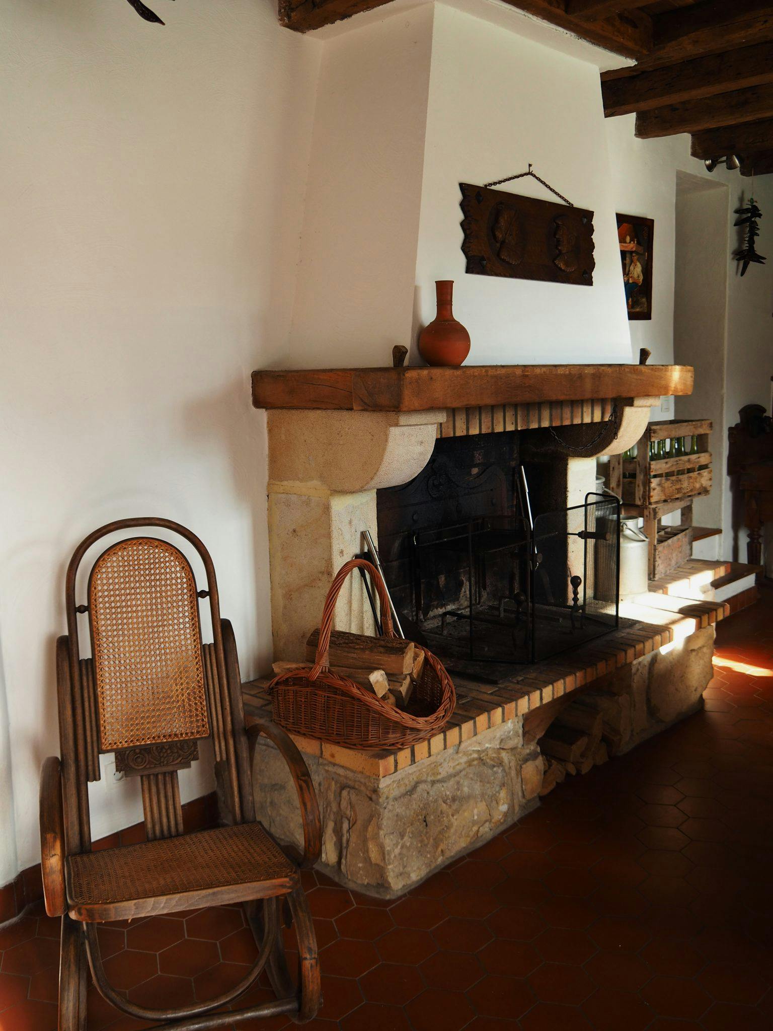 A cozy bedroom features a rustic stone fireplace with a wooden mantel. A woven chair and a wicker basket filled with logs sit nearby. Decorative pieces are hung above the fireplace. Sunlight streams in, illuminating the terracotta tiles on the floor.