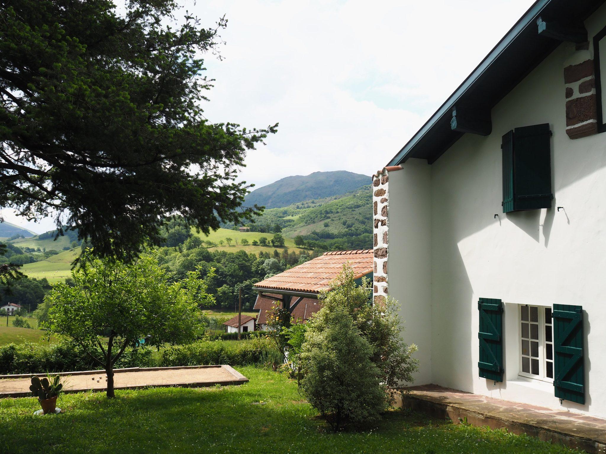 Maison aux murs blancs et volets verts dans un cadre champêtre. Un arbre et divers arbustes se trouvent dans le jardin, tandis que des collines et des montagnes sont visibles en arrière-plan. La scène est ensoleillée avec des zones nuageuses.