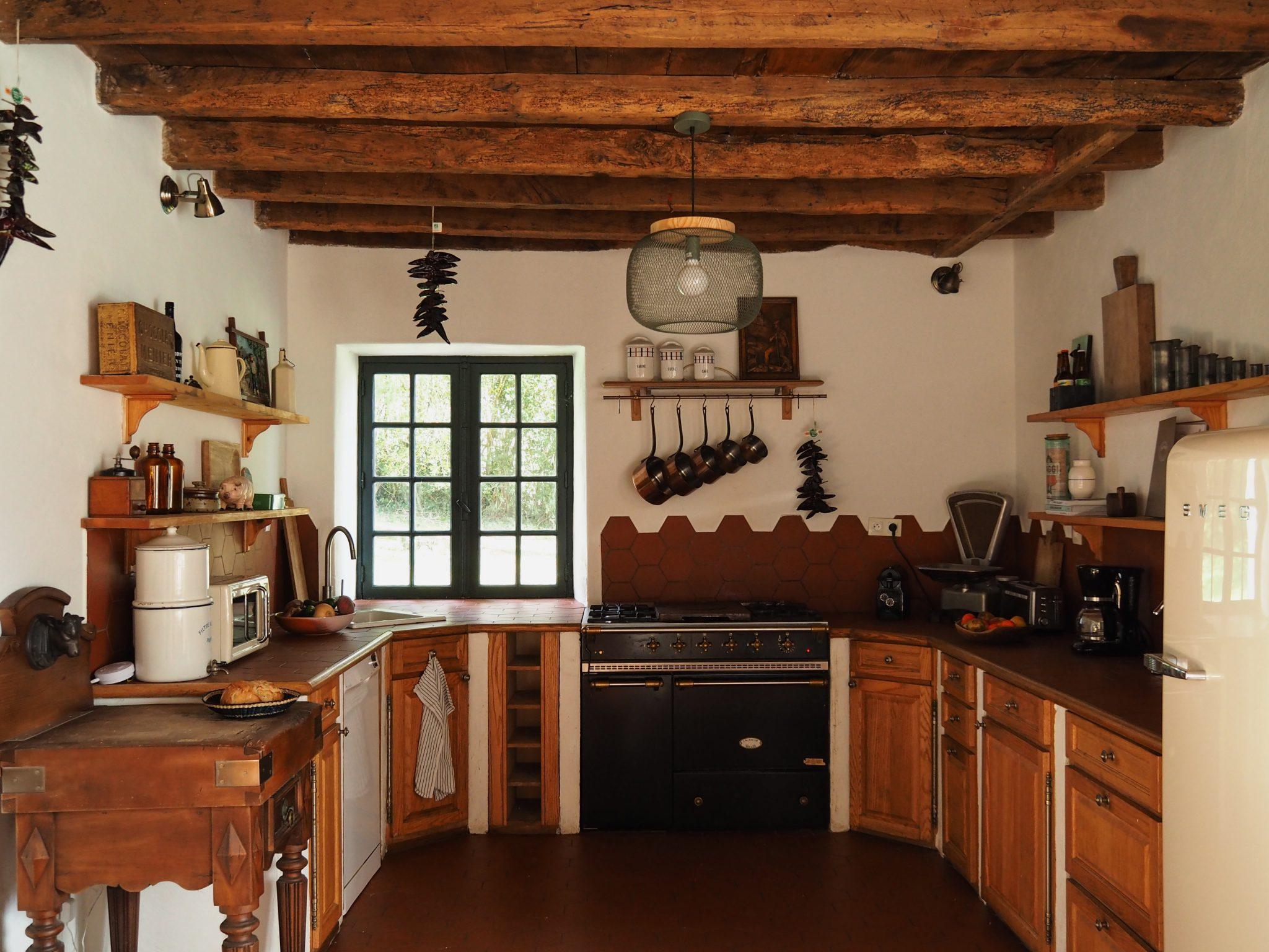 A rustic kitchen with a wooden ceiling featuring exposed beams, a large black stove, wooden cabinets, a small window, dried peppers hanging, open shelves with various kitchen items, and a retro-style refrigerator. The kitchen exudes a cozy and vintage charm.