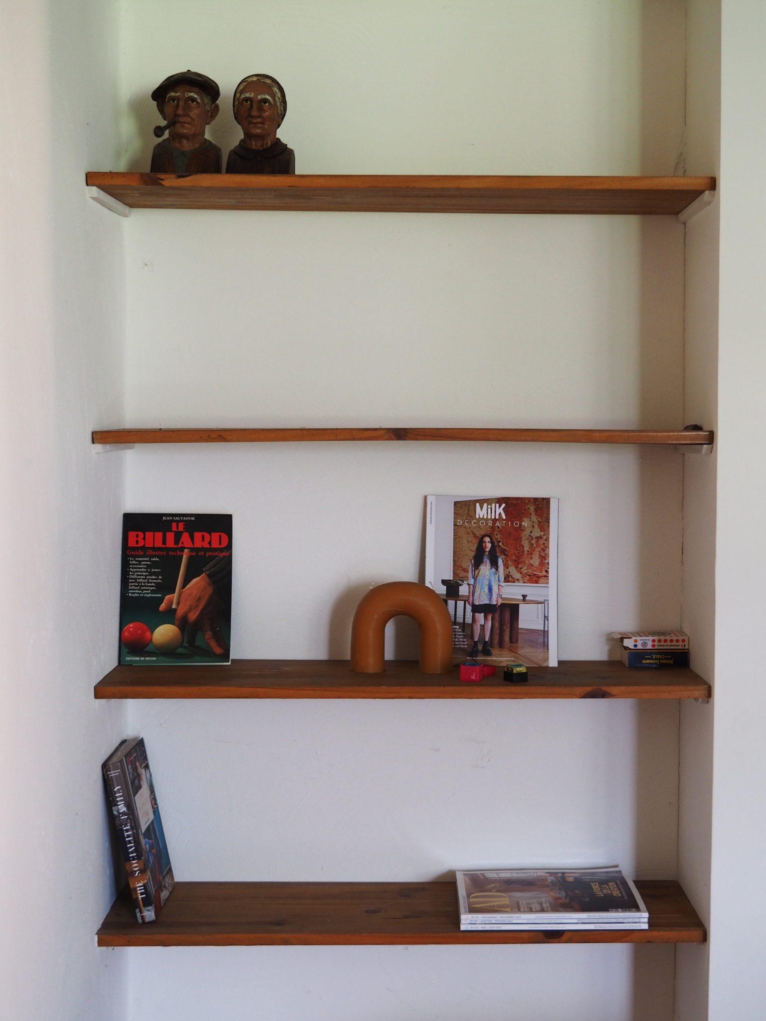 A set of wooden wall shelves in a corner displays a variety of objects. The top shelf holds two wooden busts. The middle shelves house a billiards magazine, a fashion magazine titled "Milk," and small decorative items. The bottom shelf contains a stack of books and magazines.