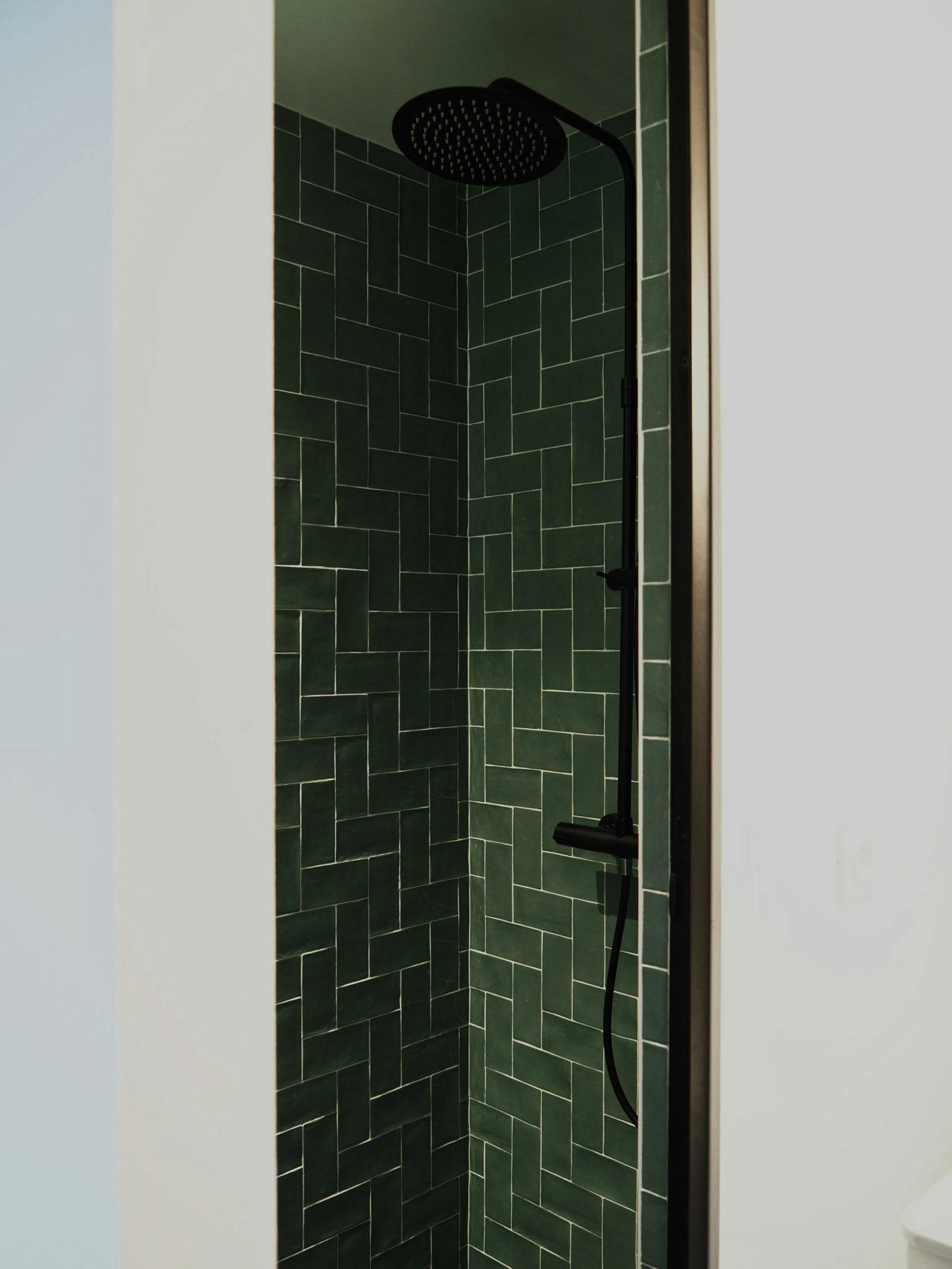 A modern shower with dark green herringbone tiles and a black rain showerhead. The shower area is partially enclosed by light-colored walls, giving it a clean and elegant look.