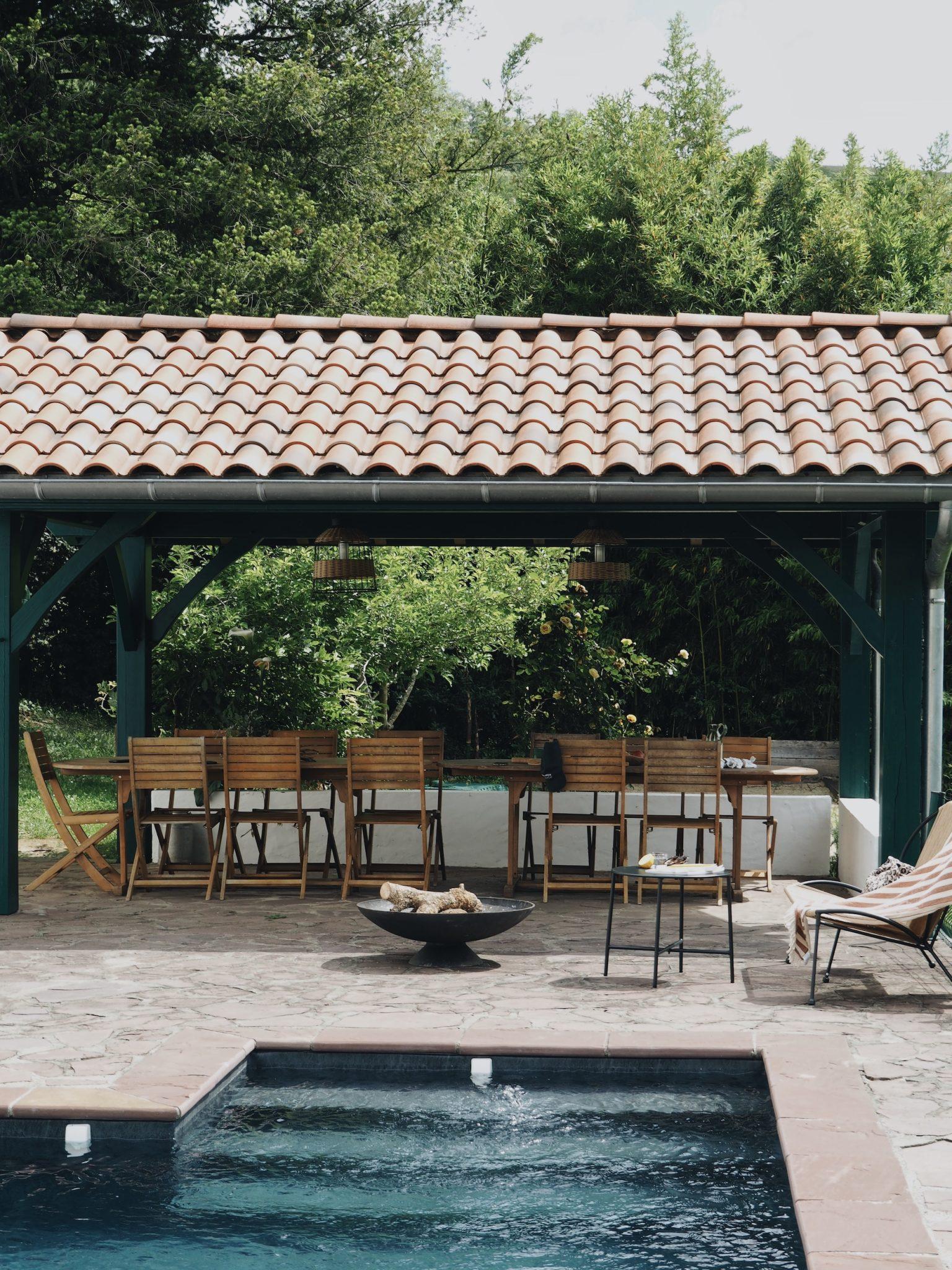 A serene outdoor space featuring a covered terrace with a tiled roof, a wooden table and chairs, all surrounded by lush greenery. In the foreground, a pool and a circular fire pit with seating contribute to the tranquil setting.