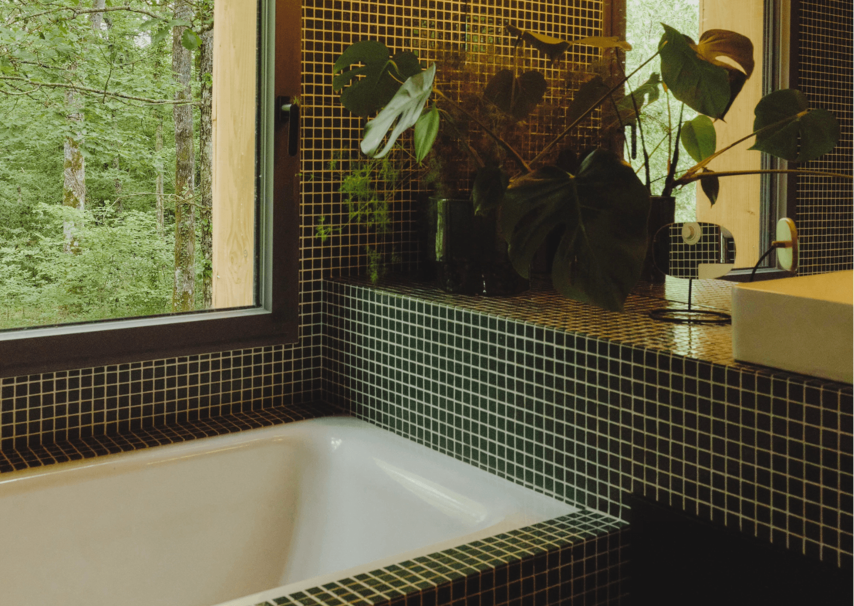 A bathroom with dark green mosaic tiles features a white bathtub. A large window next to the bathtub offers a view of a lush, verdant forest. Embracing the philosophy of Maison Sauvage, several large leafy plants are placed on the countertop, adding a touch of nature to the interior space.