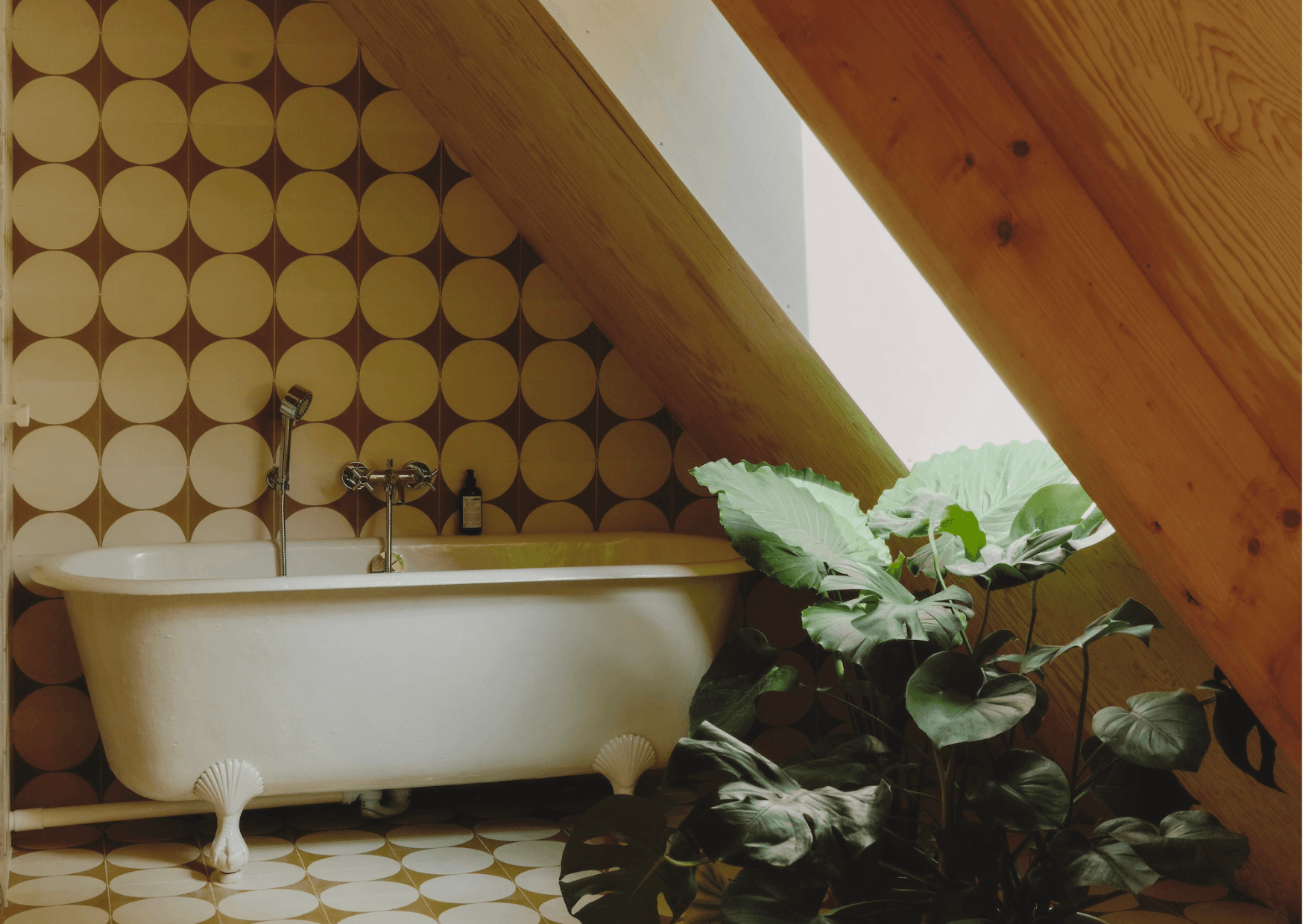 A cozy bathroom from Maison Sauvage features a white freestanding bathtub with clawfoot details, positioned against a retro circular-patterned wall. A slanted wooden skylight allows natural light to pour in, illuminating a large leafy plant next to the bathtub and enhancing the earthy ambiance.